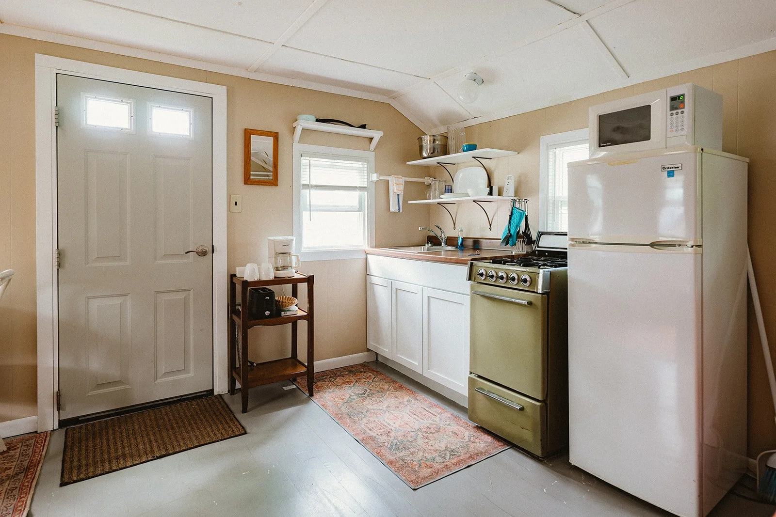 Small kitchen with white cabinetry, a stove, a refrigerator with a microwave on top, open shelves, a coffee maker, and two windows.