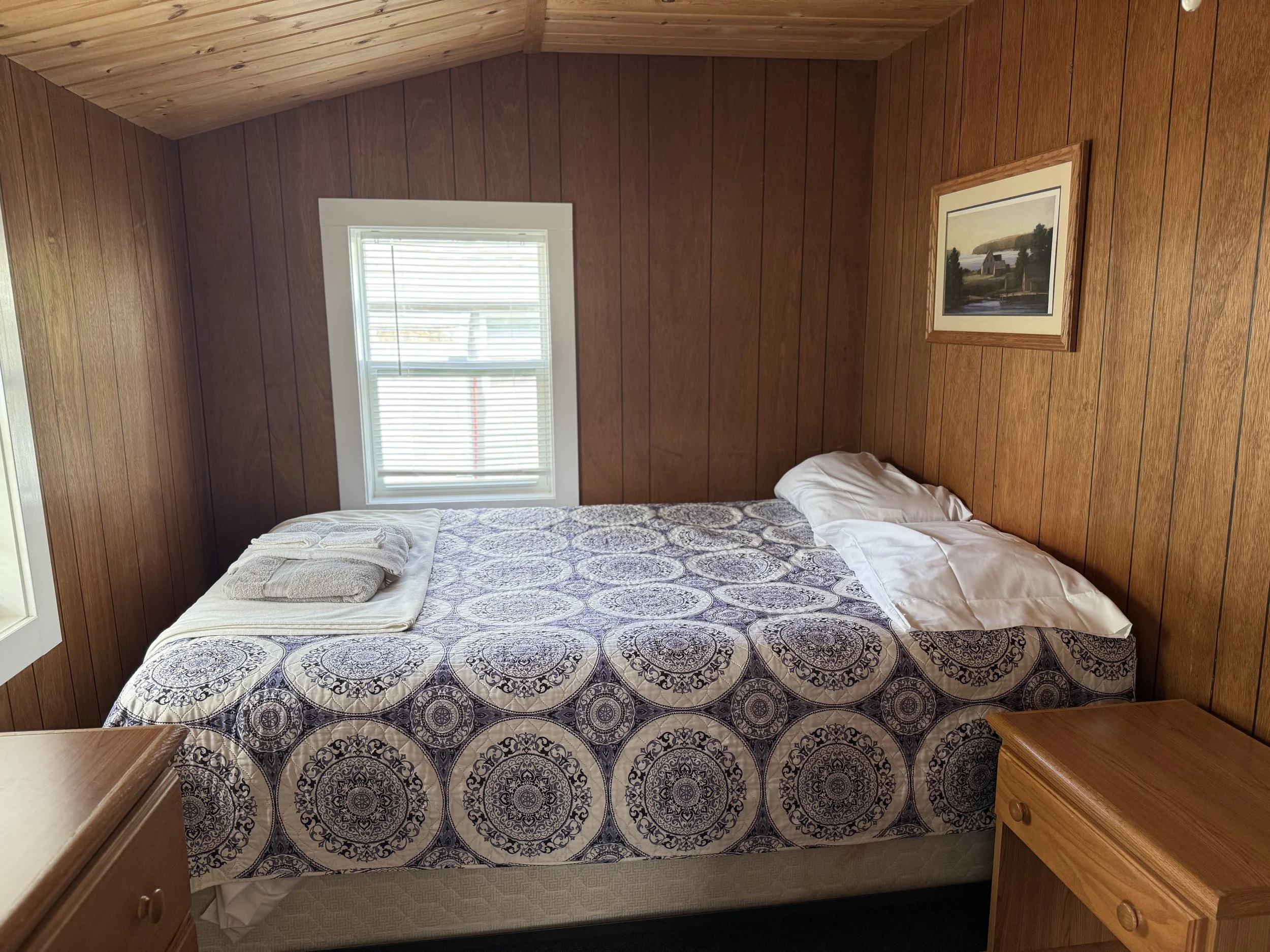 Bedroom with wooden walls, a window with blinds, a bed with patterned bedding, pillows, and towels, and a framed landscape painting on the wall. Brookside Lake Cottages Duck Lake, Grawn, MI