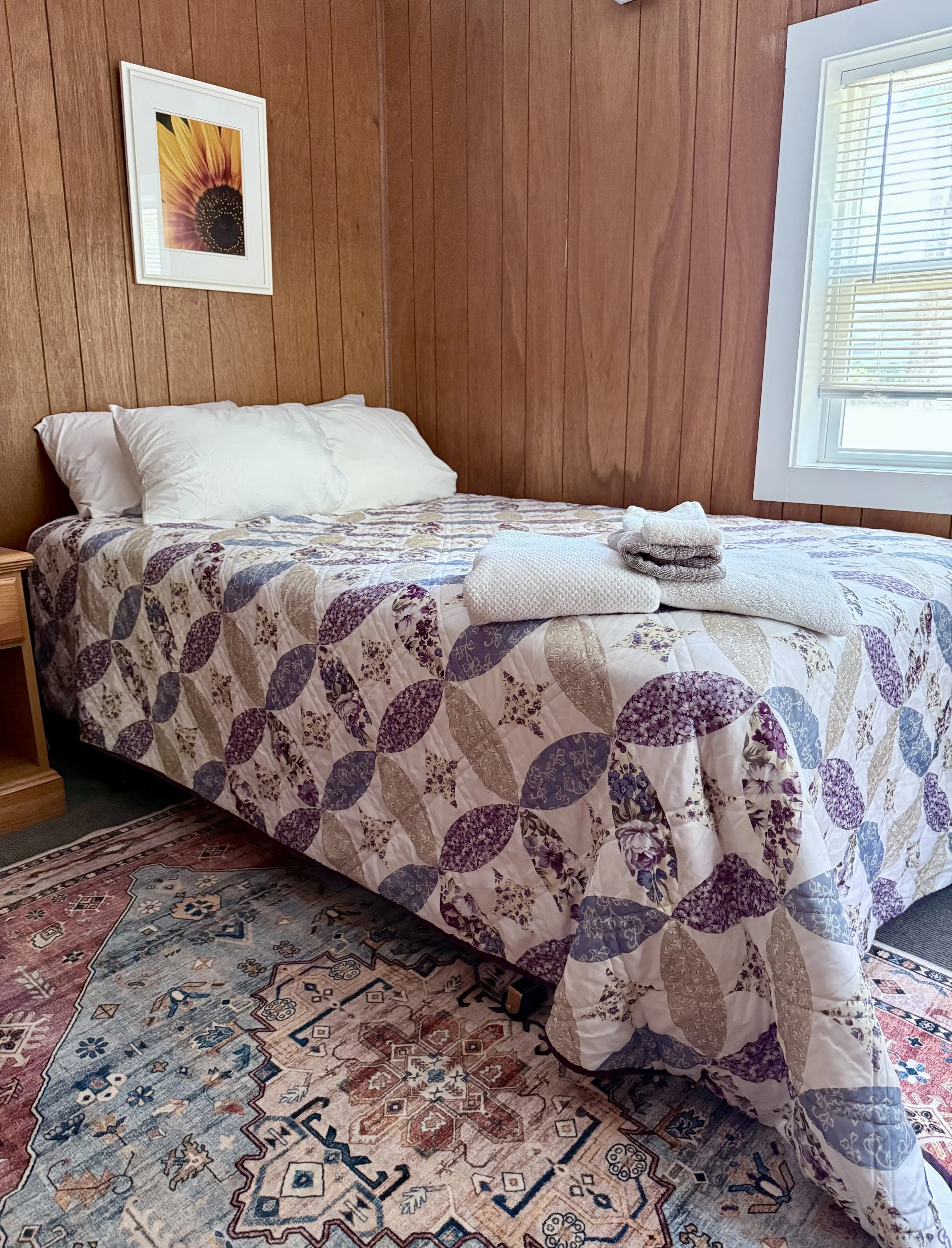 A bedroom with wood-paneled walls, a bed with a patterned quilt, white pillows, and folded towels on top. There is a window with blinds and a colorful framed flower picture on the wall.