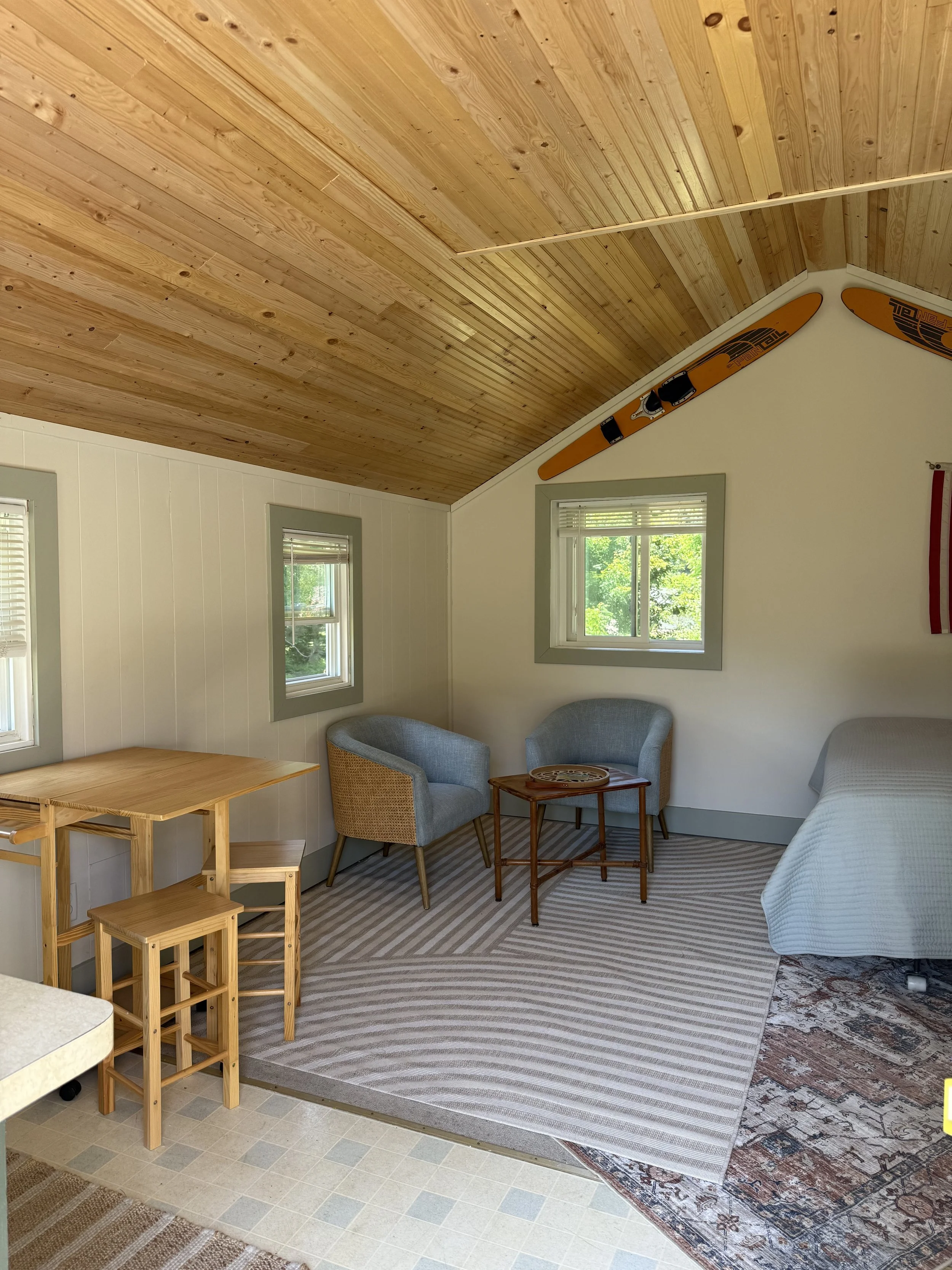 A cozy room with a sloped wooden ceiling, two small windows, two light blue armchairs, a small wooden table, and a bed with a light-colored cover. There is a wood-paneled wall, a striped rug, and skis mounted on the wall.