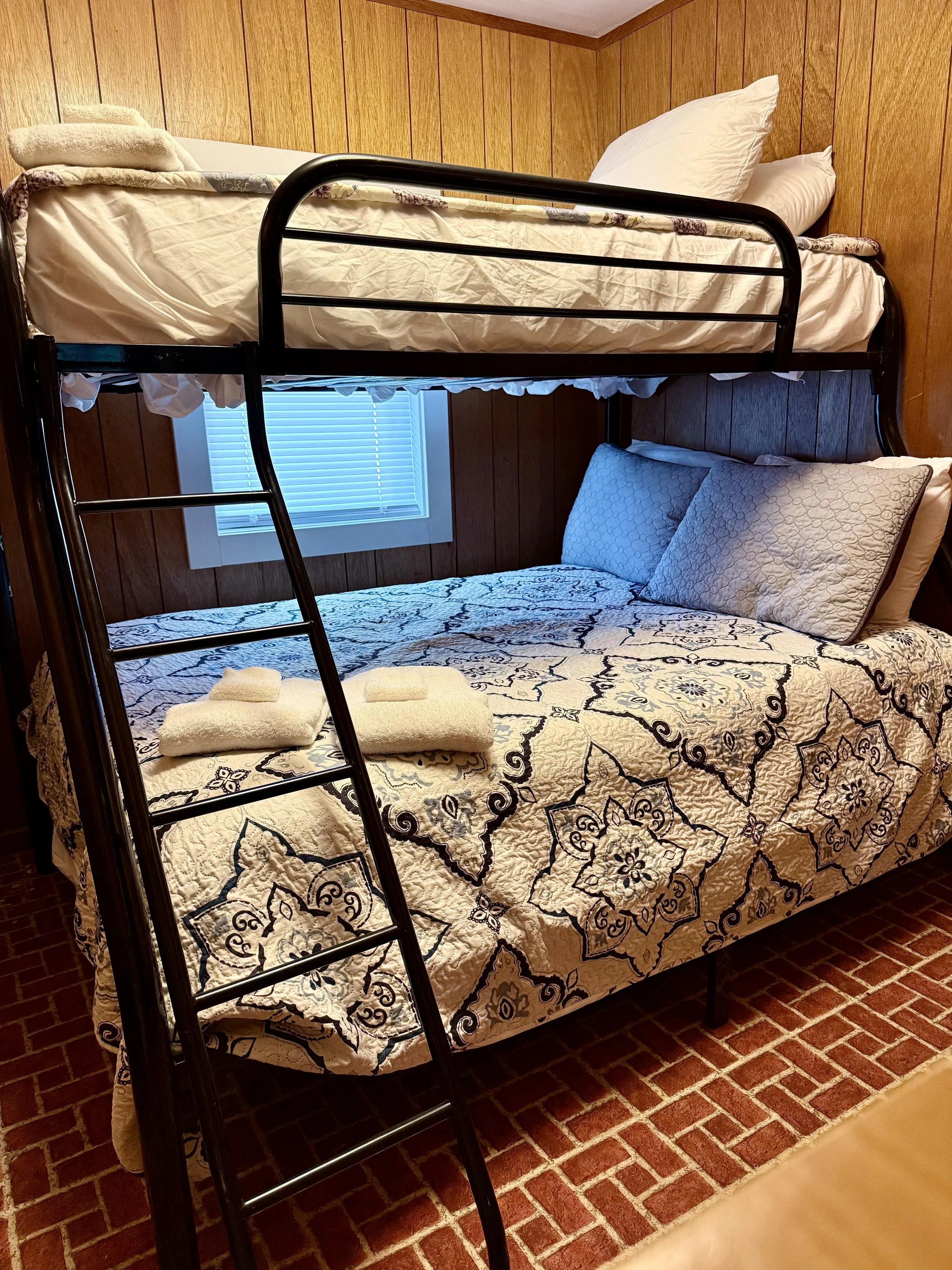 A bunk bed with a triple-mattress bed on the bottom and a twin-size bed on top, with pillows and towels, in a room with wood-paneled walls and a window with blinds.
