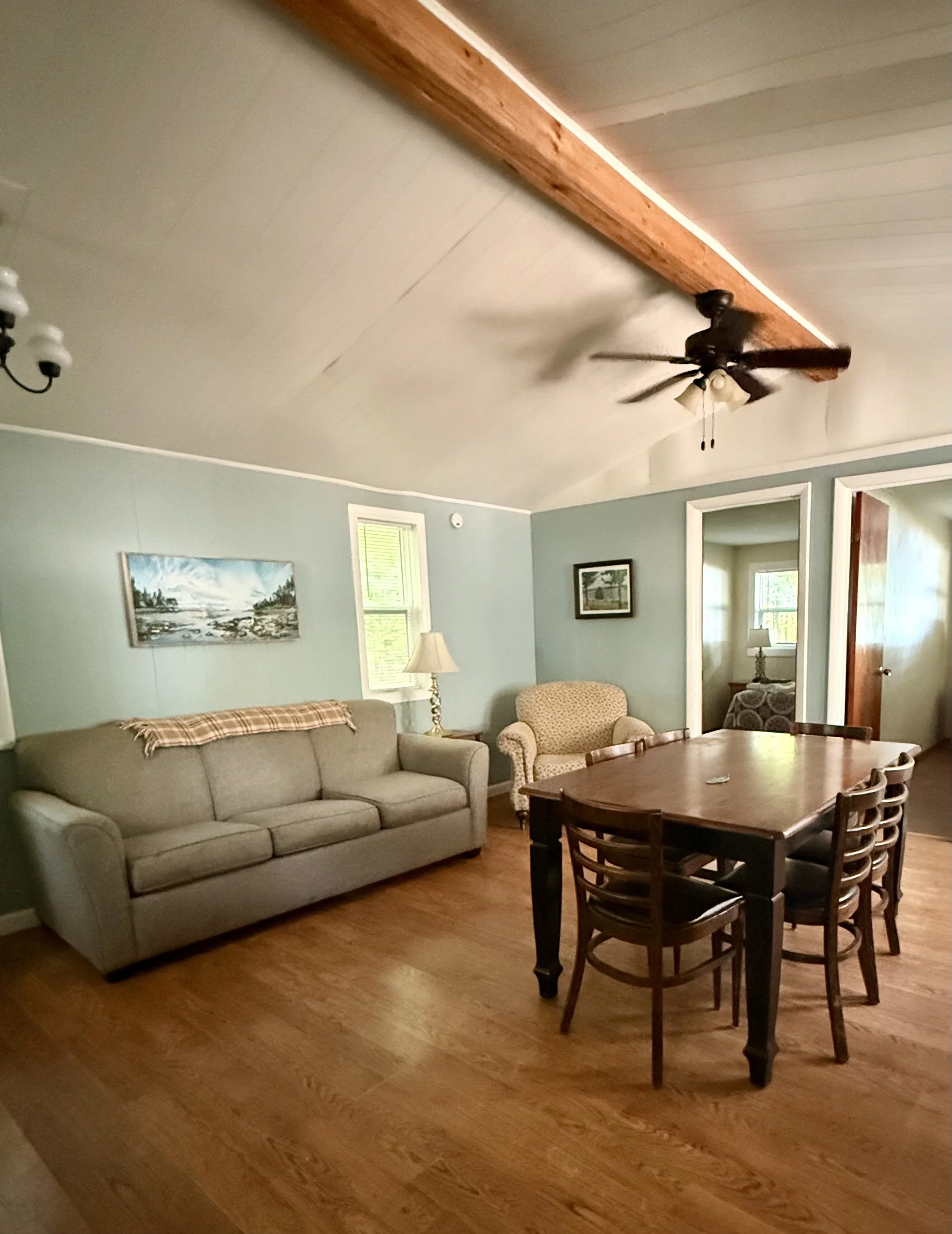 Living room with a beige sofa, an armchair, a wooden dining table with chairs, hardwood floor, a ceiling fan, and paintings on the walls. Brookside Lake Cottages Duck Lake, Grawn, MI