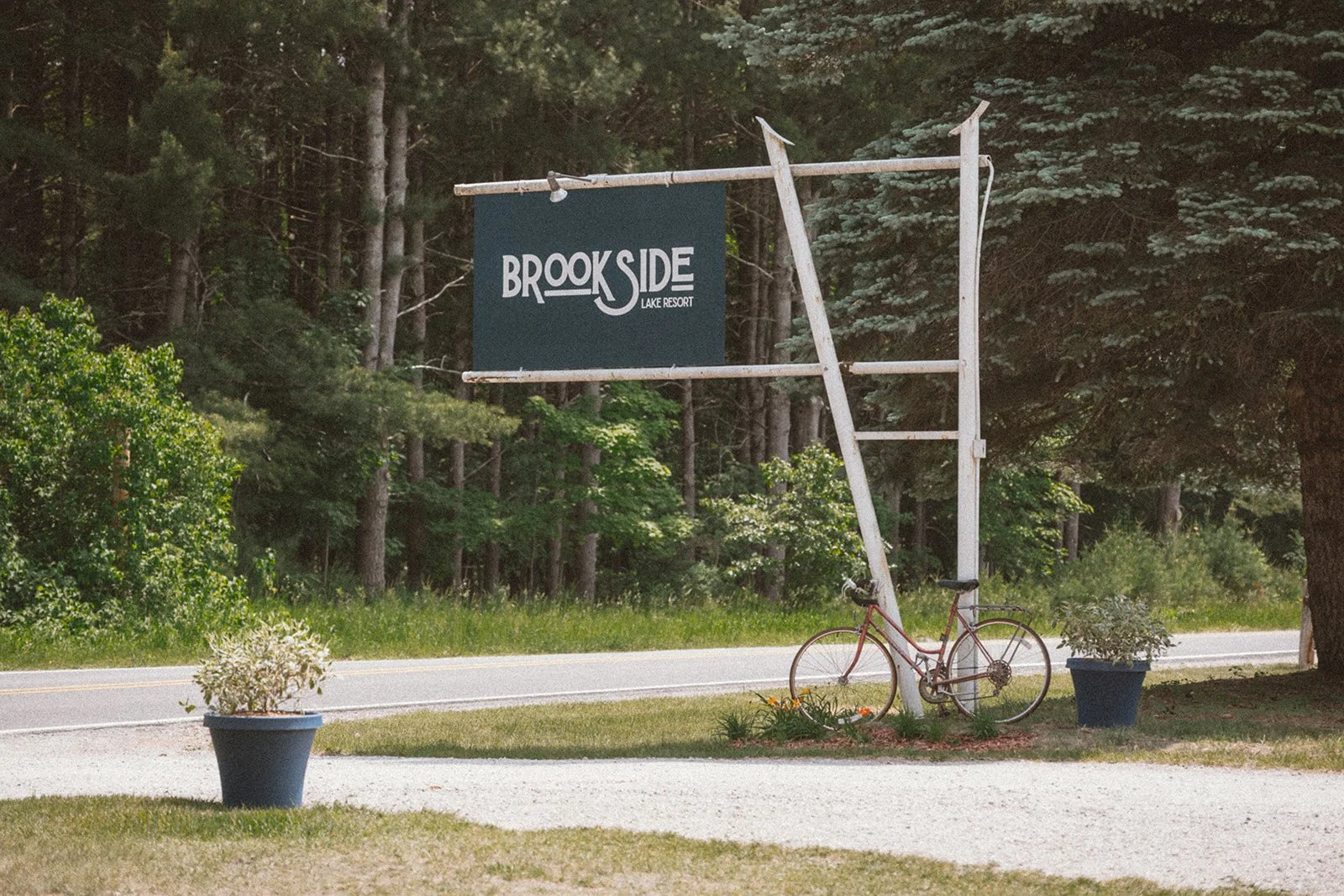 A sign that reads 'Brookside Lake Resort' is mounted on a wooden frame beside a road. There is a red bicycle leaning against the frame and two large planters with green plants are on either side of the sign. Trees and greenery are in the background.
