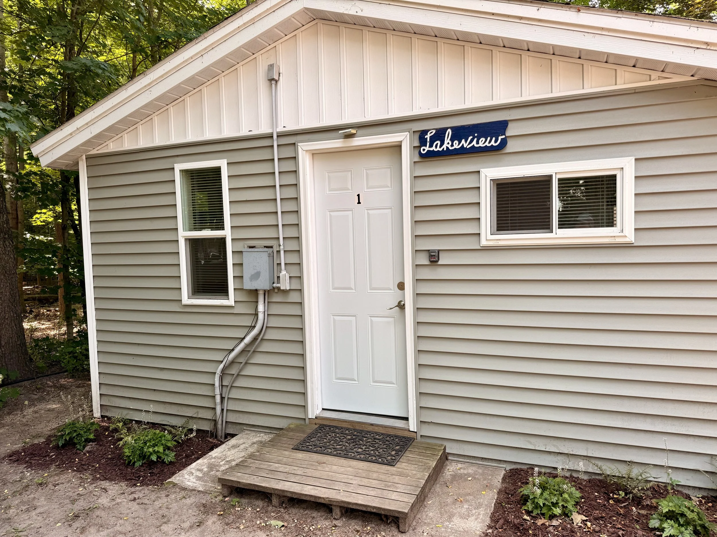 Exterior of a beige house with a white door labeled '1' in a small front yard with plants, a wooden step, and a sign that says 'Lakesview' above the door.