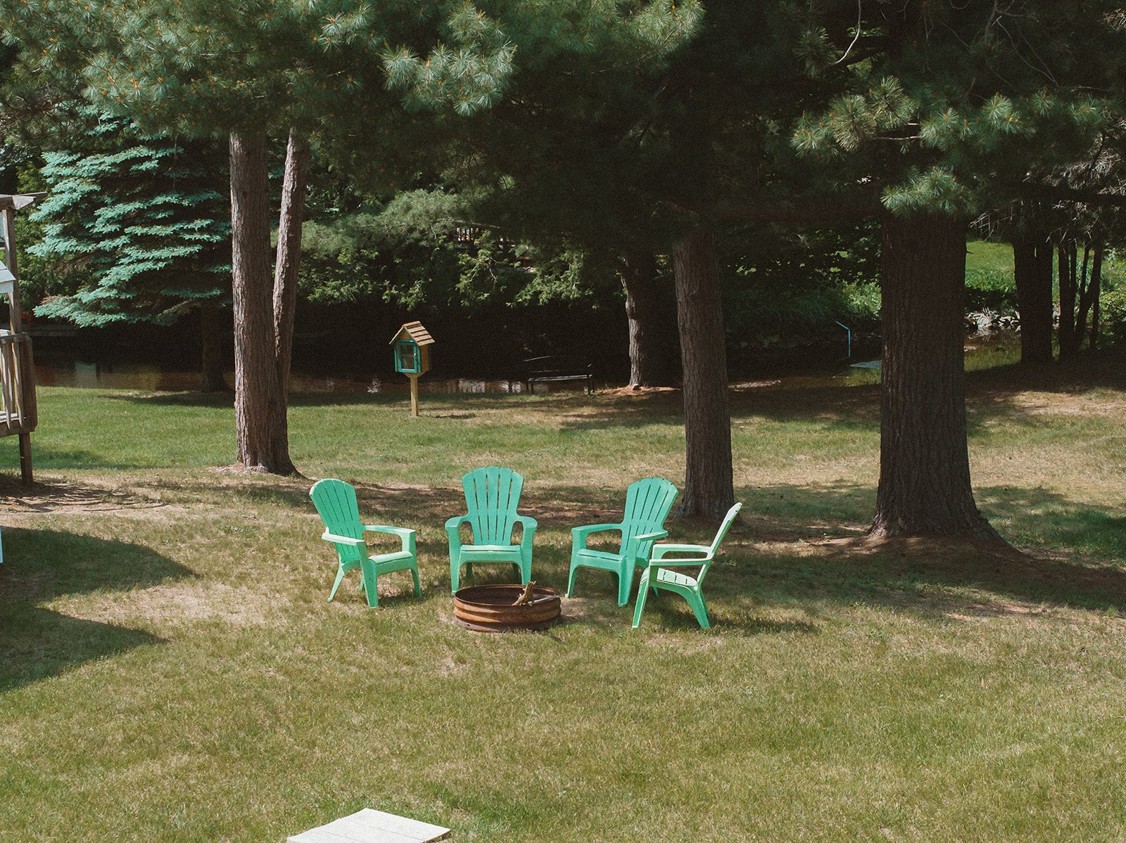 Four turquoise lawn chairs are arranged in a circle on a grassy yard, with a firepit in the middle and tall pine trees in the background.