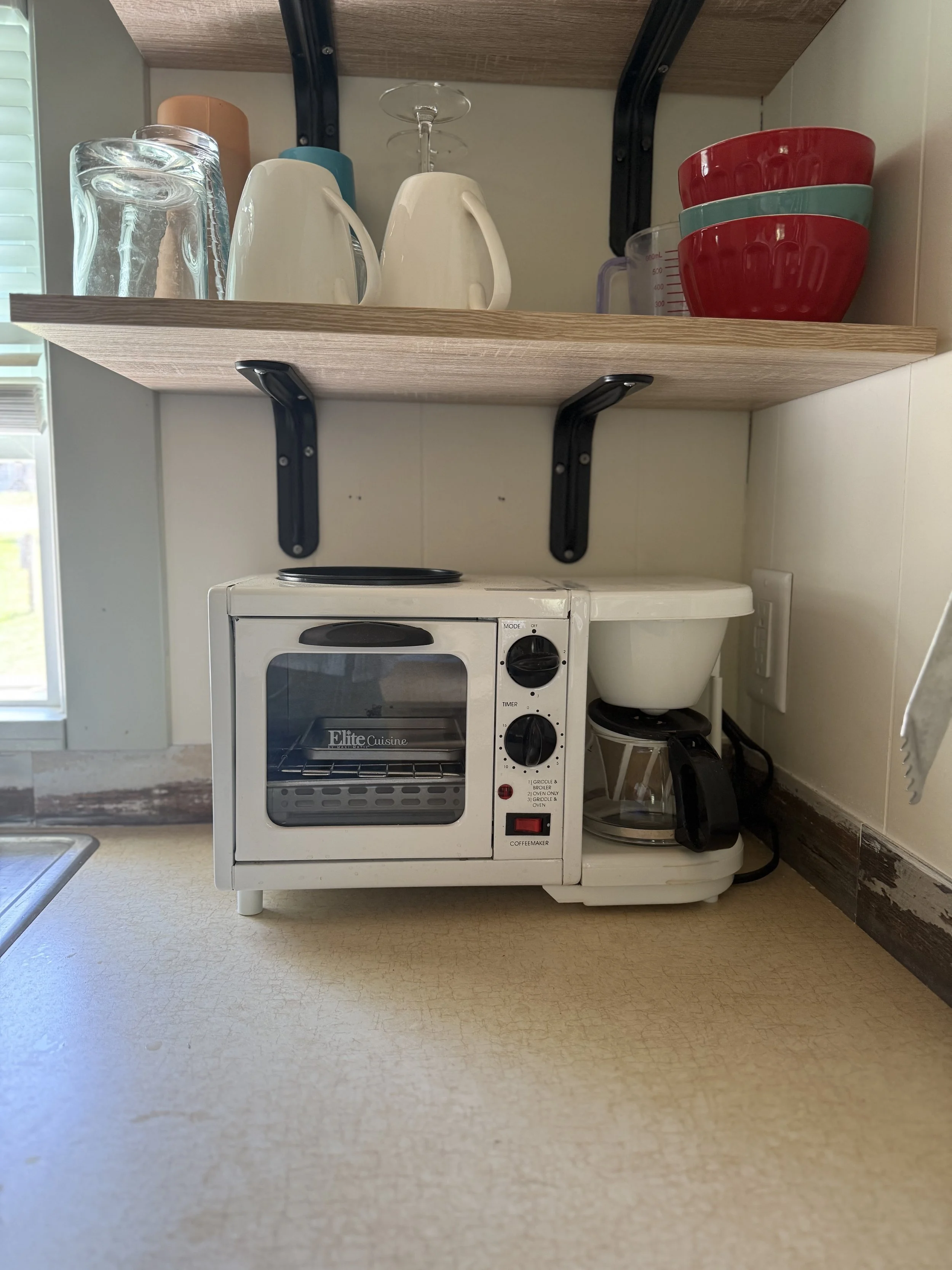 Kitchen countertop with a small white toaster oven, a coffee maker, and a white bowl. Above, a wooden shelf holds glasses, mugs, and bowls.