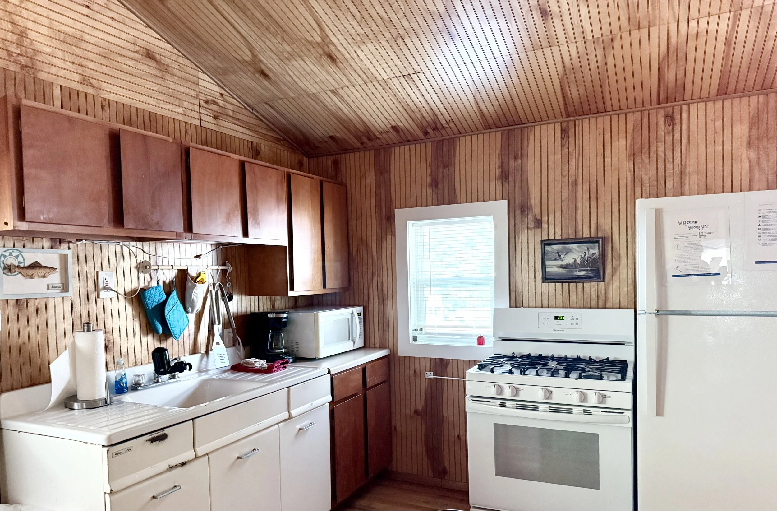 Kitchen with wooden panel walls and ceiling, white stove, refrigerator, microwave, and sink, with various kitchen items on the counter.