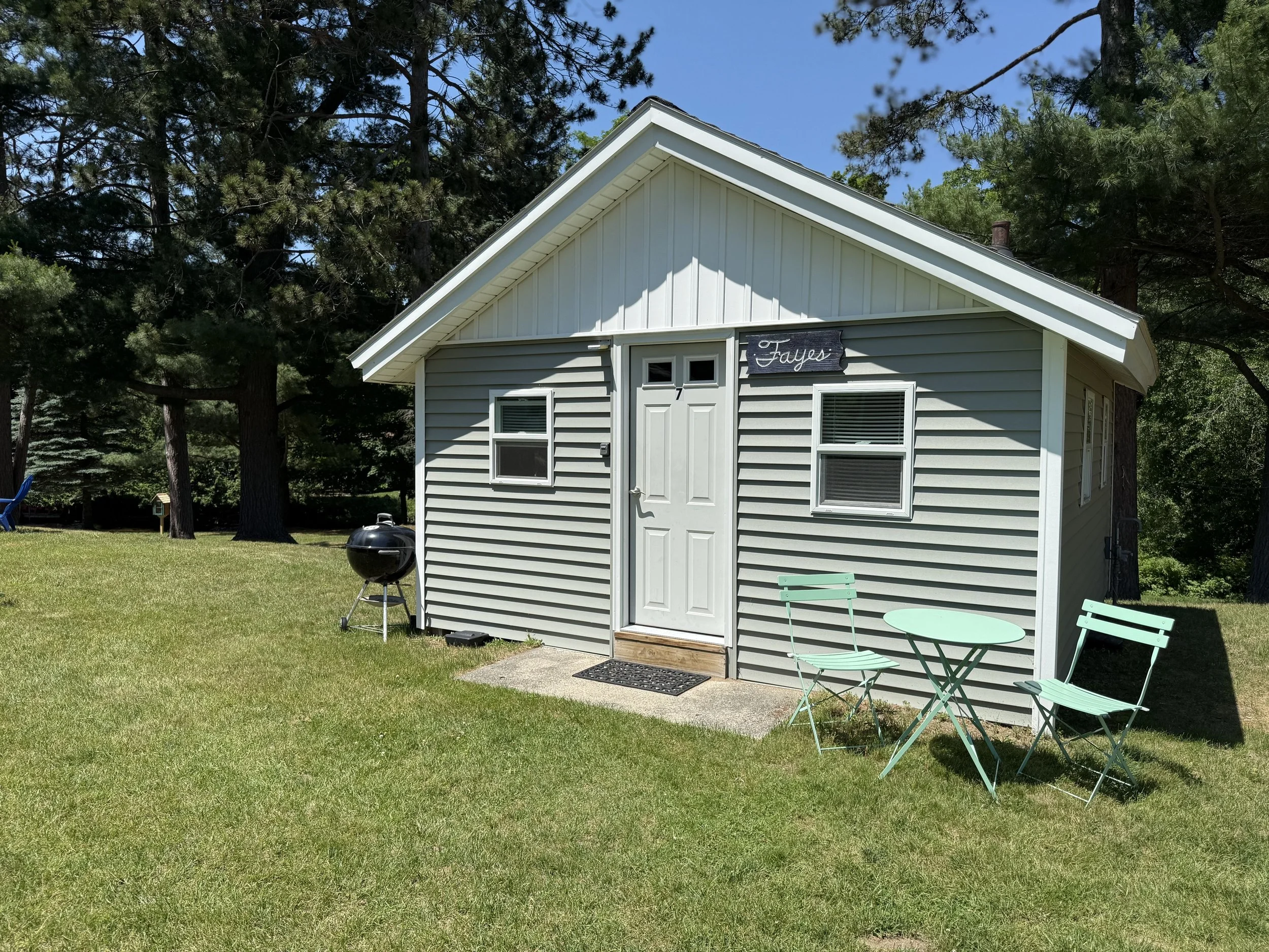 Small gray house with white trim, two windows, and a white door labeled 'Fayes' on a green lawn with trees in the background. There is a small turquoise table with two matching chairs and a black kettle grill nearby.