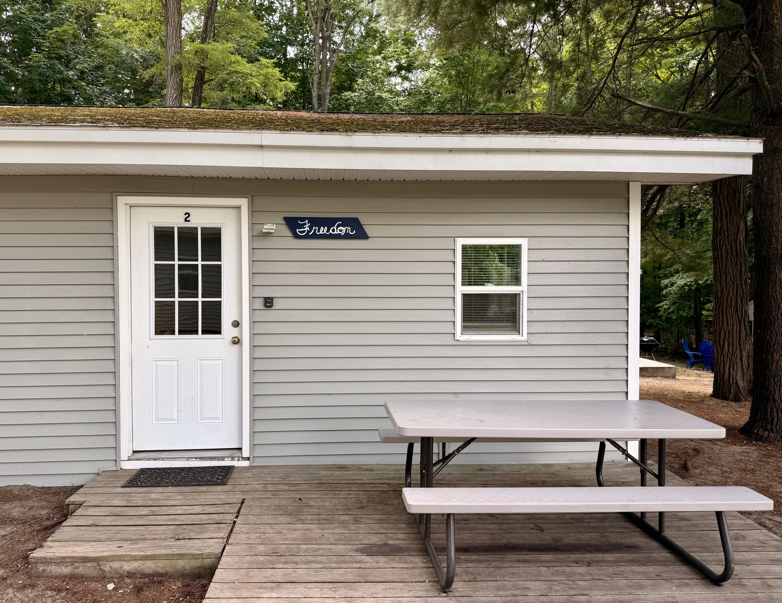 The back of a small, light gray house with a white door labeled with the number 2, a small window with blinds, and a blue sign that reads 'Freedom.' There is a wooden deck with a picnic table in front of the house, surrounded by trees and a forested