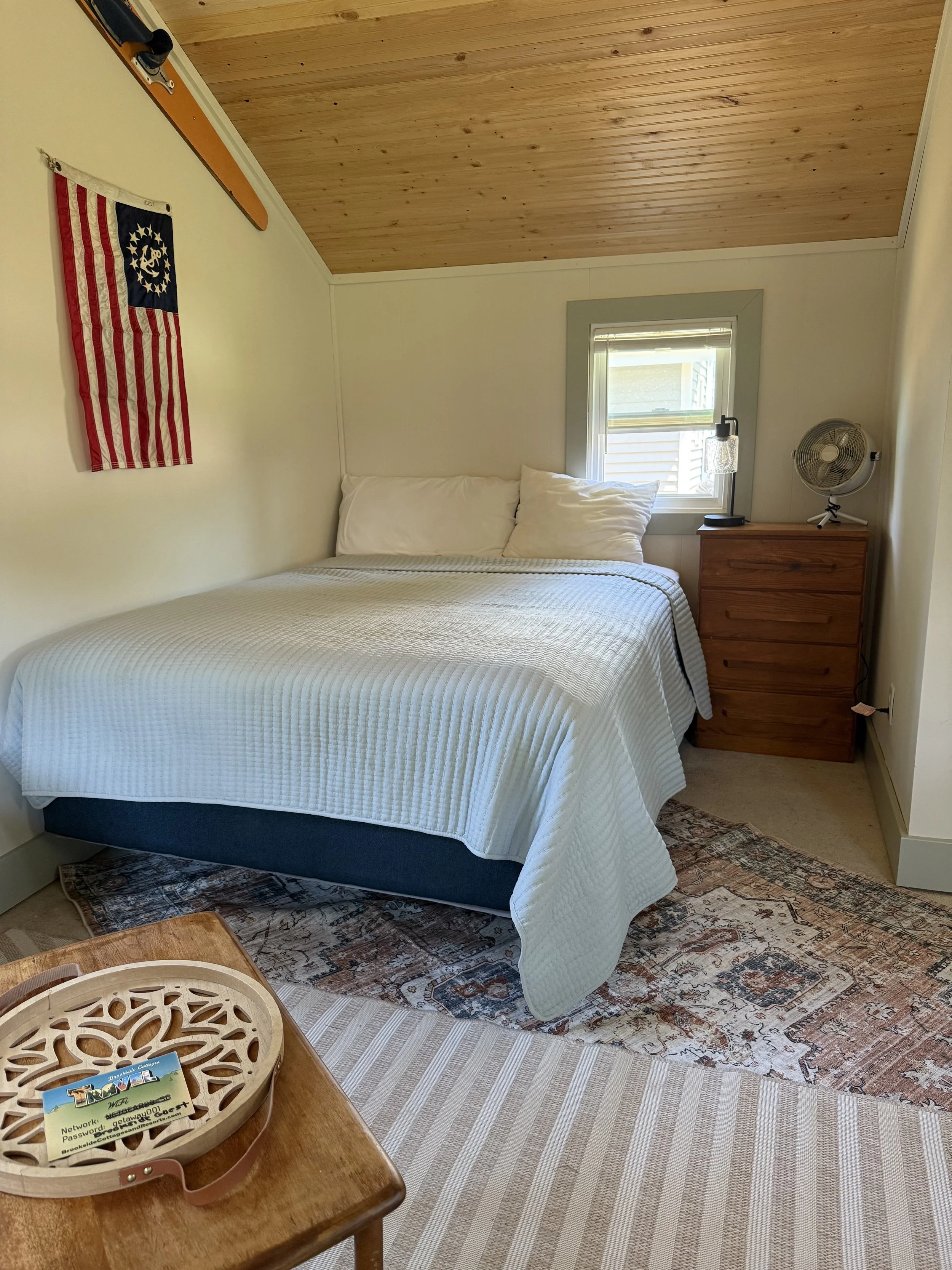 A cozy bedroom with a wooden ceiling, a window with a small lamp and fan on a wooden dresser, a bed with white bedding, a small American flag on the wall, and a tray with lanyards on a wooden table.