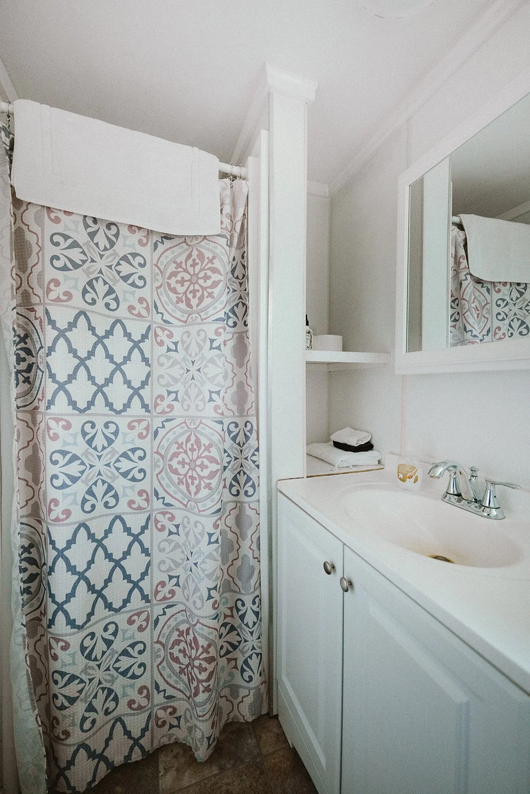 Bathroom with a shower curtain featuring a geometric pattern, a white sink with a silver faucet, a mirror, and shelves with towels and toiletries.