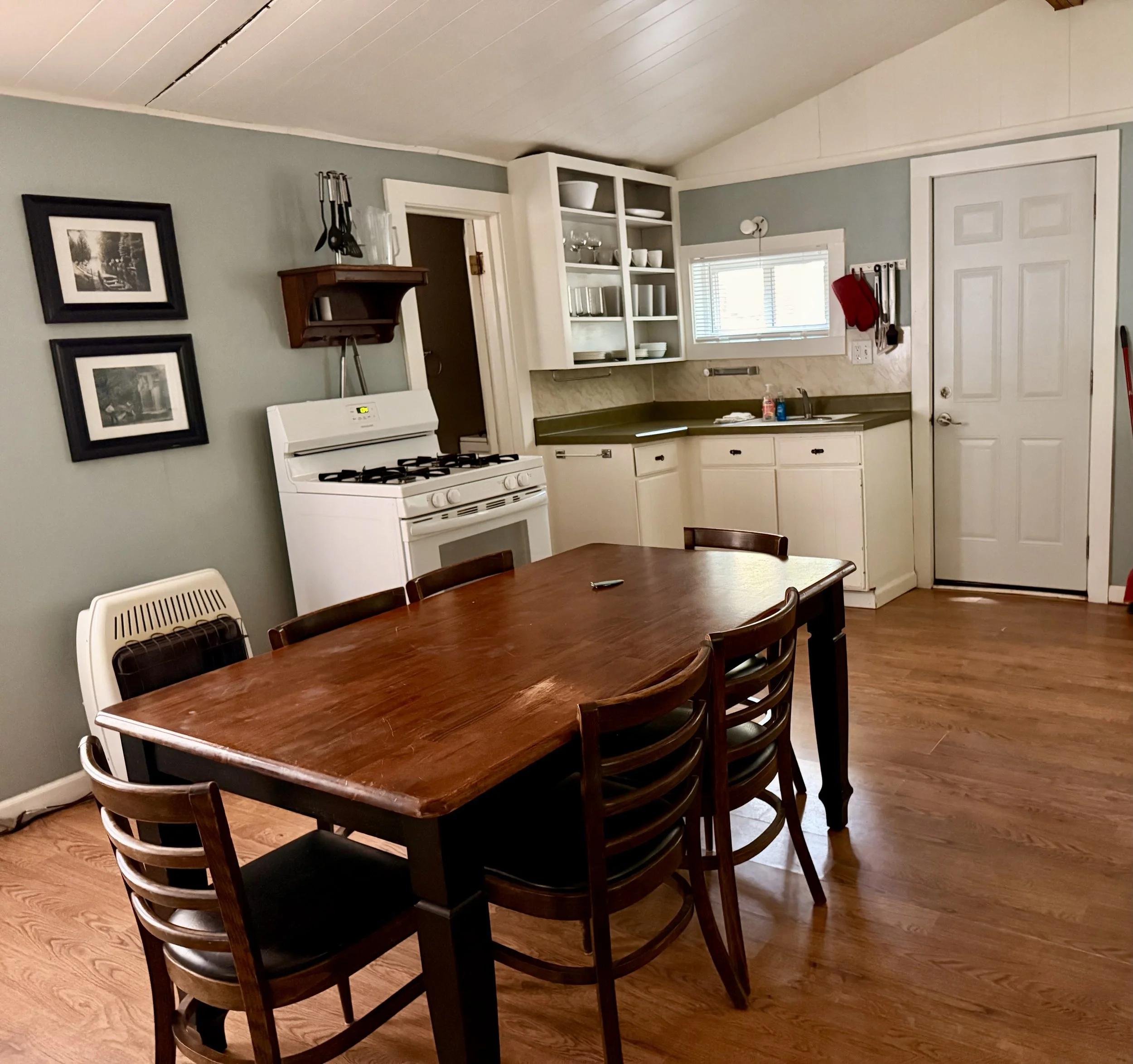 A kitchen and dining area with a wooden dining table and six chairs, a white stove, open white cabinets with dishes, a window, and a door, with a wood floor and painted walls.