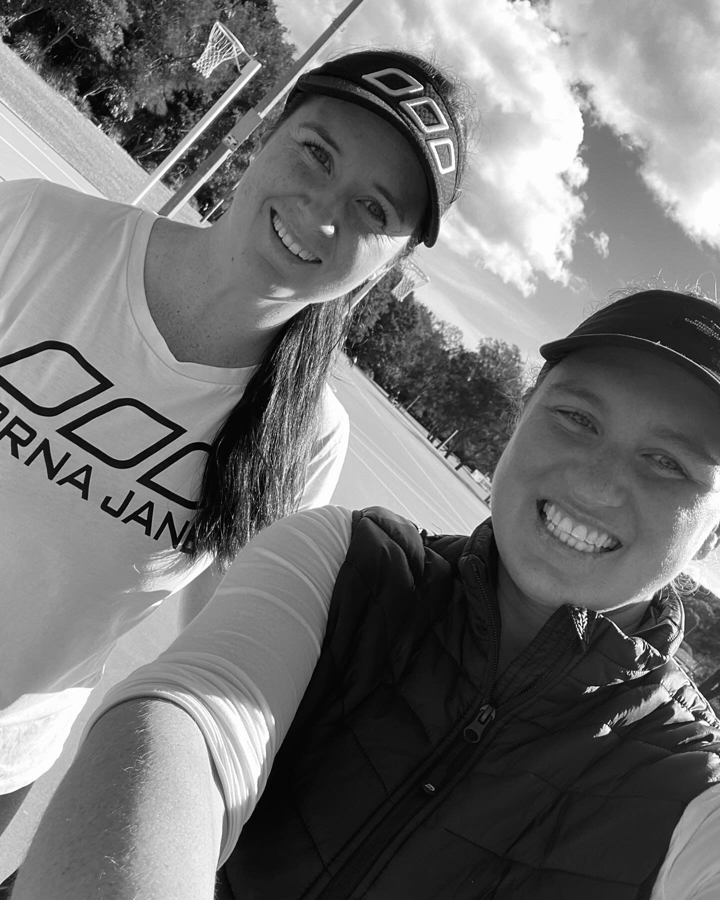 Two women smiling for a selfie outdoors on a sunny day, with a basketball hoop and trees in the background.