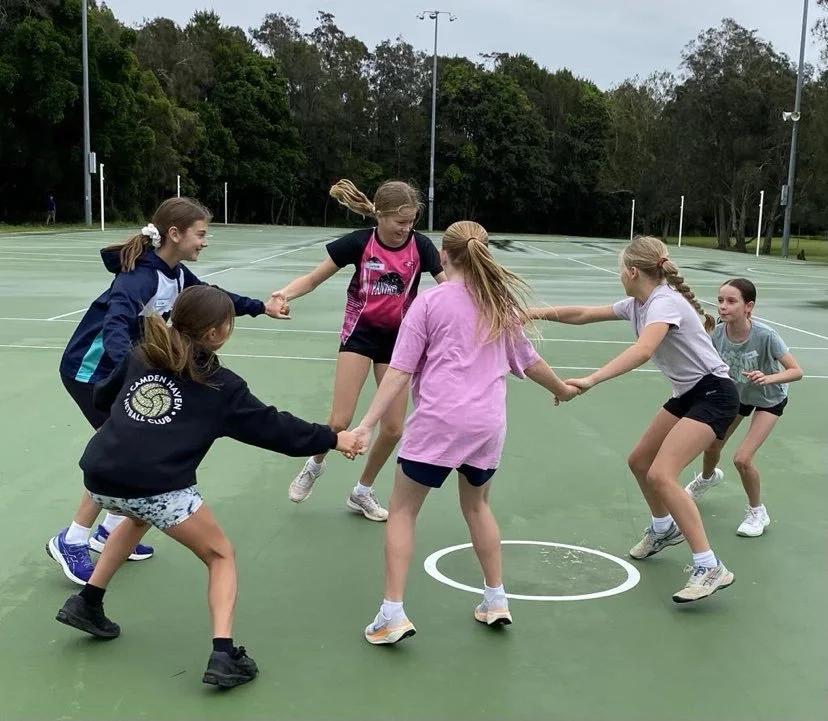 Six young girls are playing a game on a green outdoor basketball court, holding hands and forming a circle.