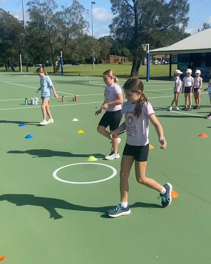 Group of children participating in outdoor activities on a green basketball court, with cones and water bottles set up, under a partly cloudy sky, in a park with trees and a pavilion in the background.