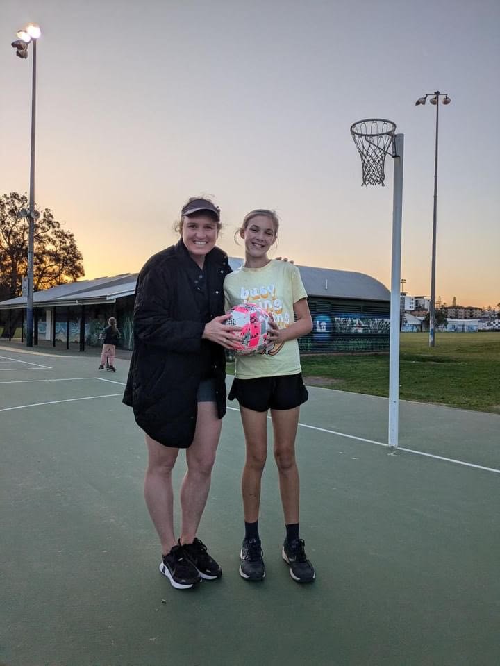 Two young women standing on a basketball court at sunset, holding a colorful basketball, smiling at the camera.