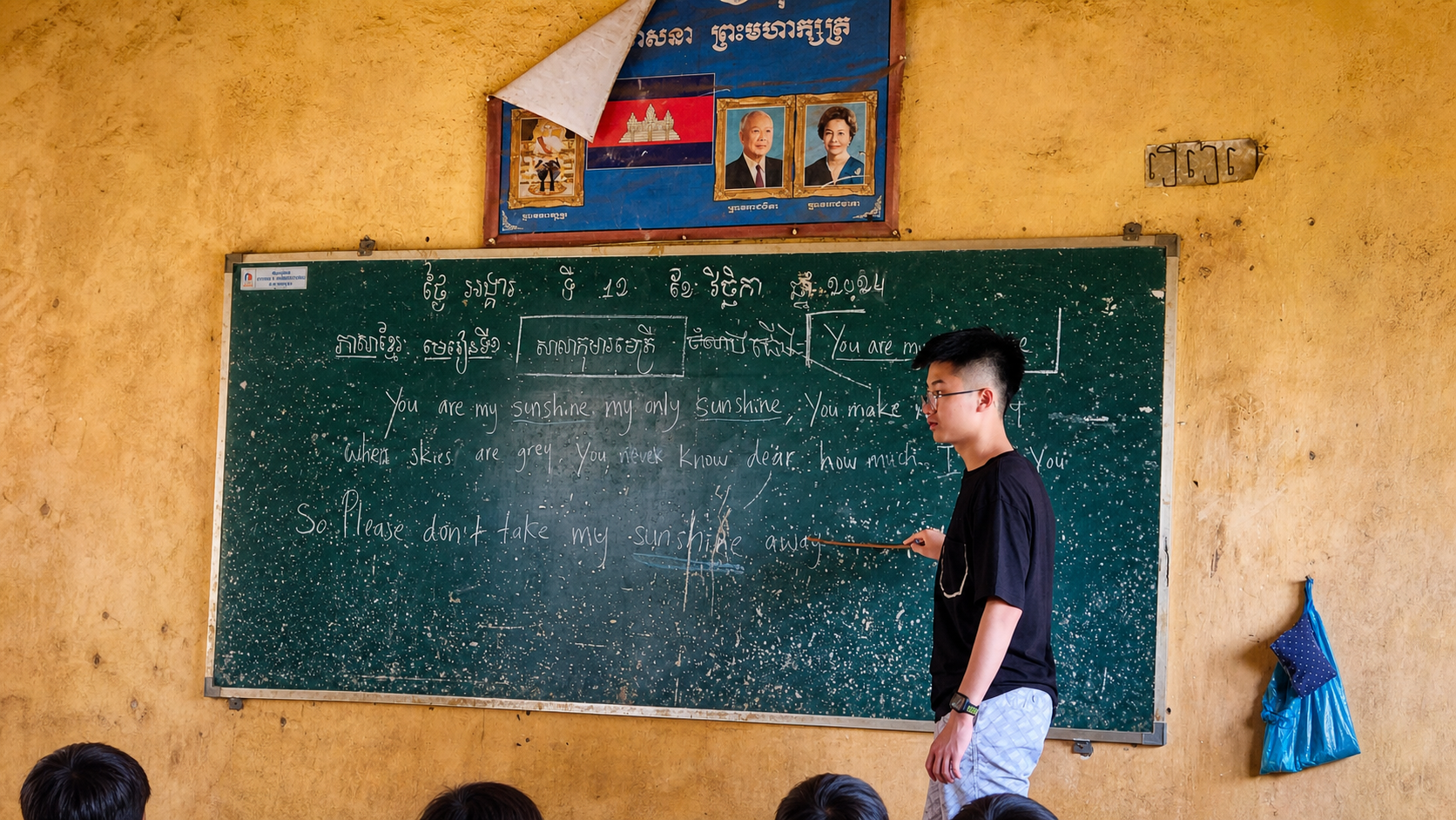 Jerome Ong pointing at English song lyrics written on a blackboard in a Cambodian village primary school.