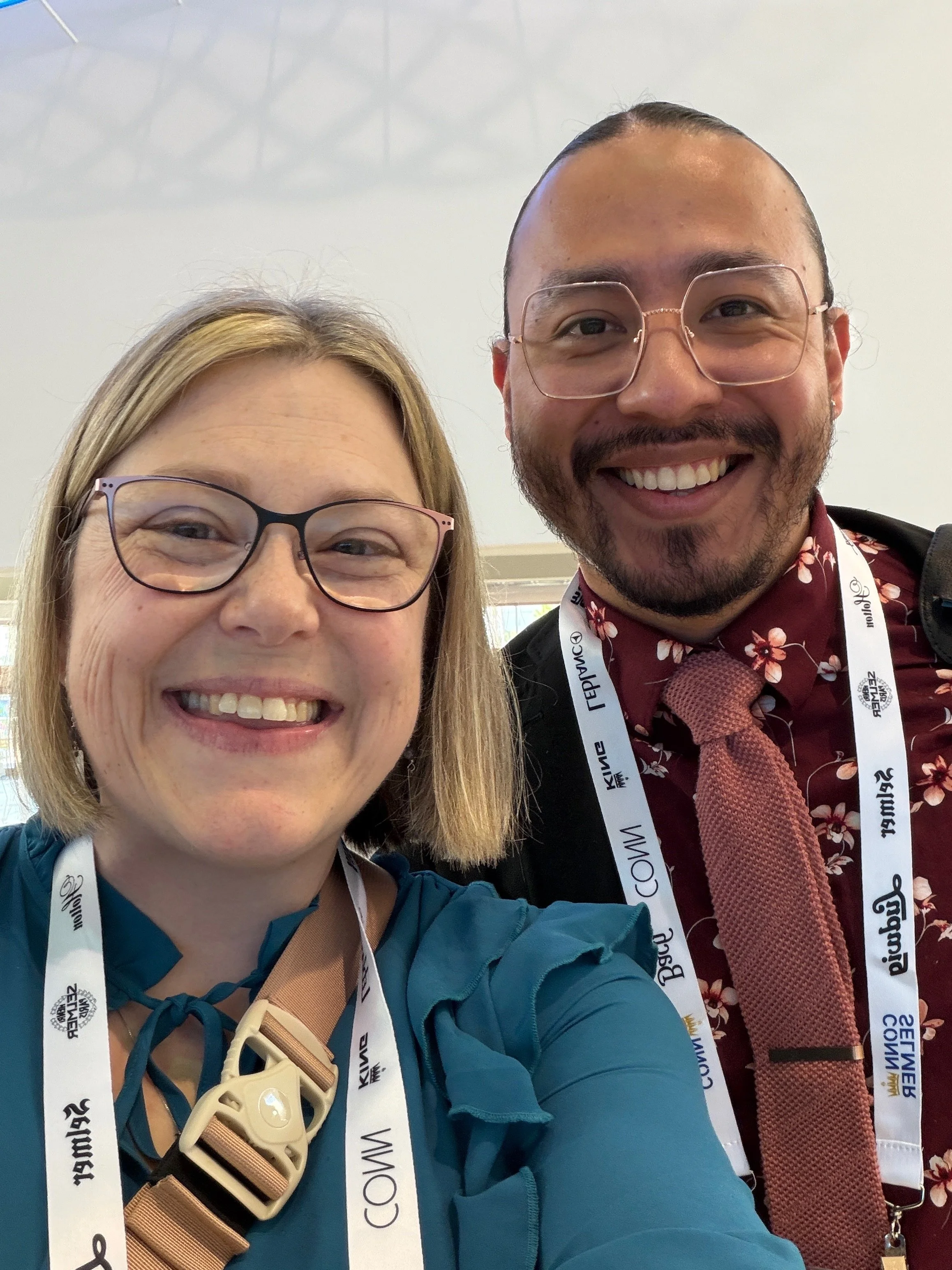 Two smiling individuals taking a selfie at a conference or event, both wearing badges and lanyards. The woman on the left has blonde hair, glasses, and is wearing a blue jacket with a beige strap across her chest. The man on the right has a beard, gl