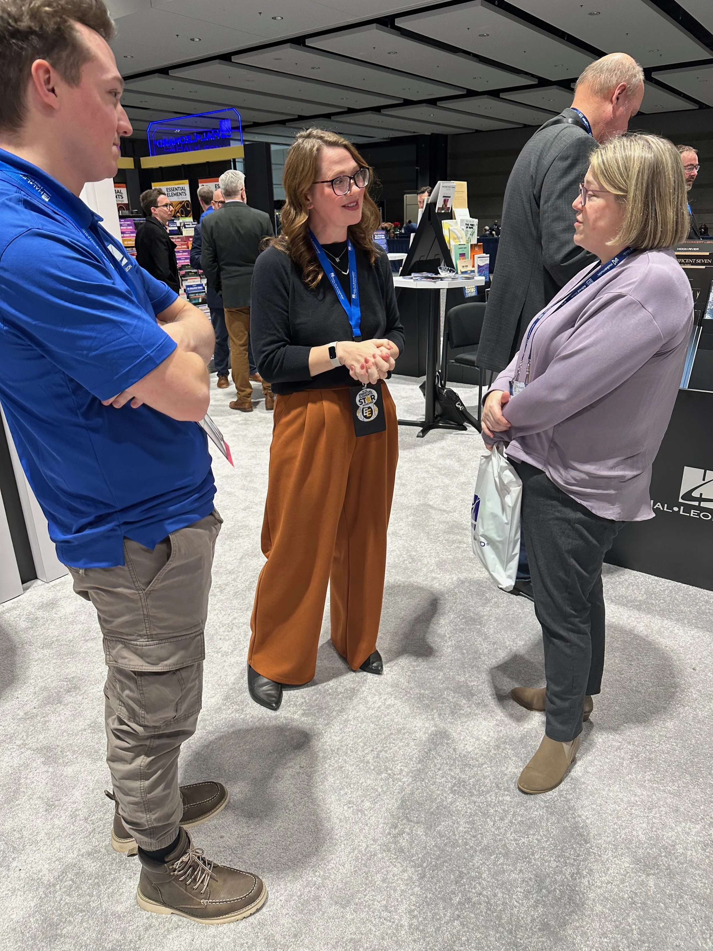 Four people engaged in conversation at a conference or convention, with books and promotional materials displayed in the background.