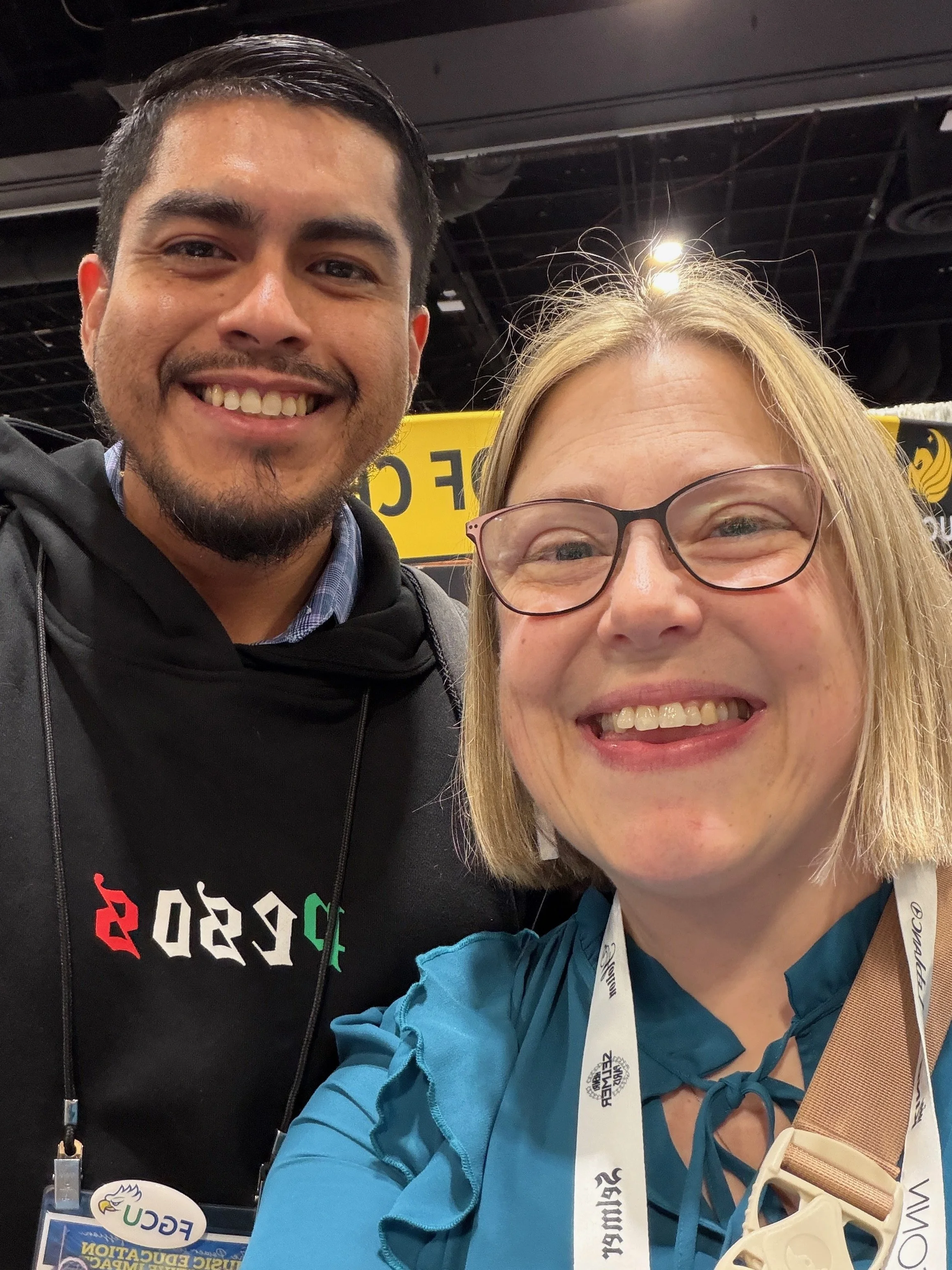 A smiling man and woman taking a selfie at an event, with badges and lanyards around their necks and a yellow banner in the background.