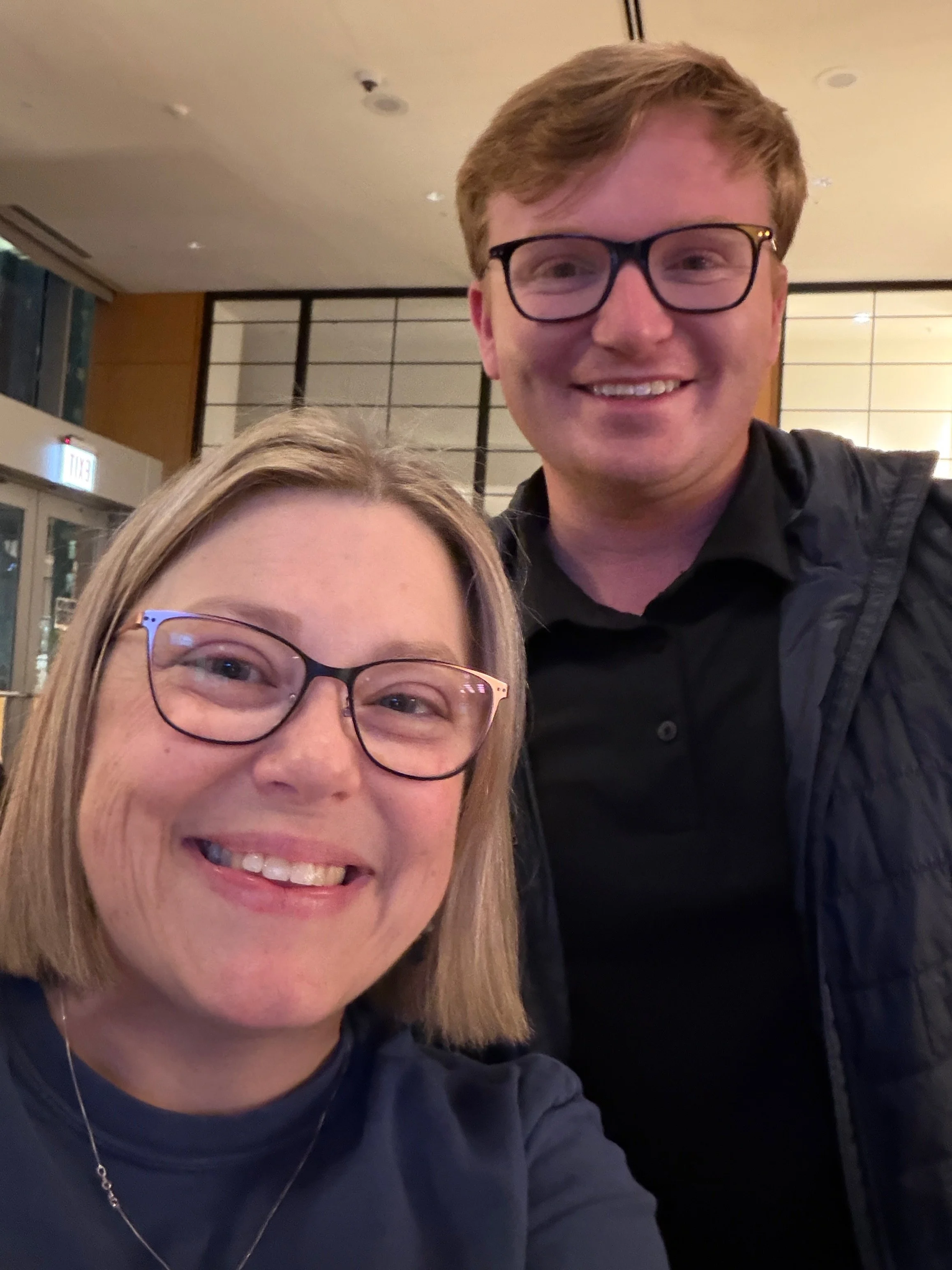 A smiling woman with glasses and blonde hair in the foreground, and a smiling man with glasses and light brown hair in the background, both taking a selfie in an indoor setting.