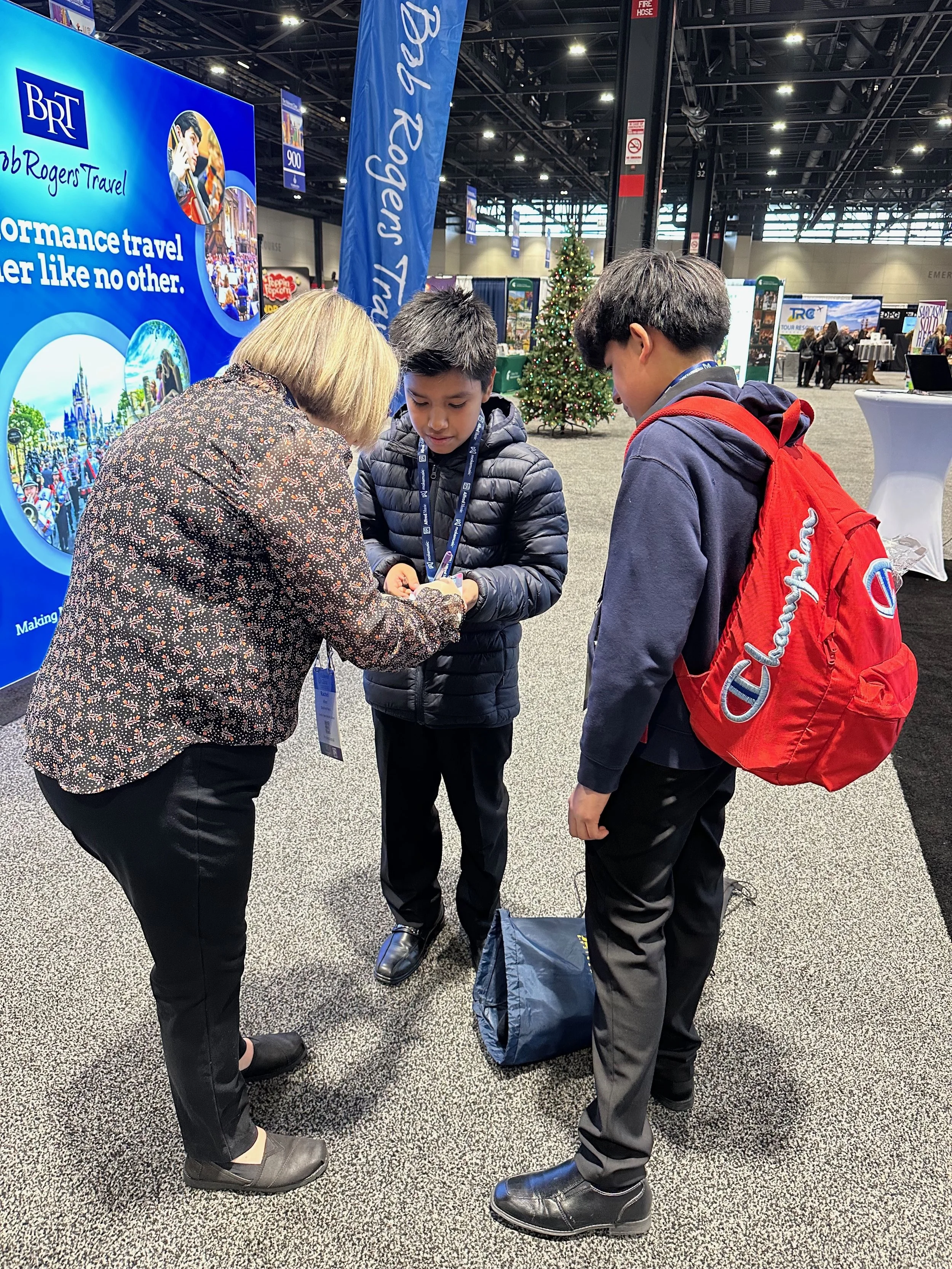 A woman and two boys standing at an indoor event, looking at an object the woman is holding. The woman has blonde hair and is dressed in a patterned blouse and black pants. One boy has short dark hair and is wearing a black puffy jacket. The other bo