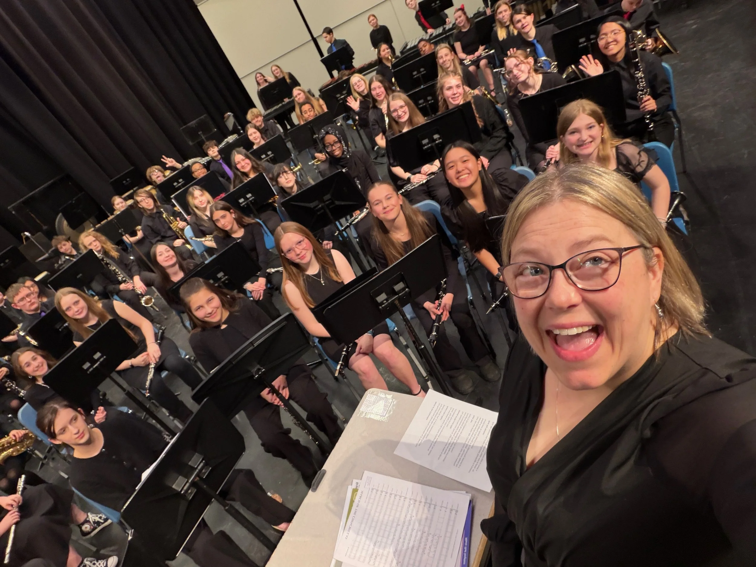 A large group of young orchestra musicians sitting with their instruments on stage, smiling and looking at the camera, with a music stand and sheet music in the foreground.