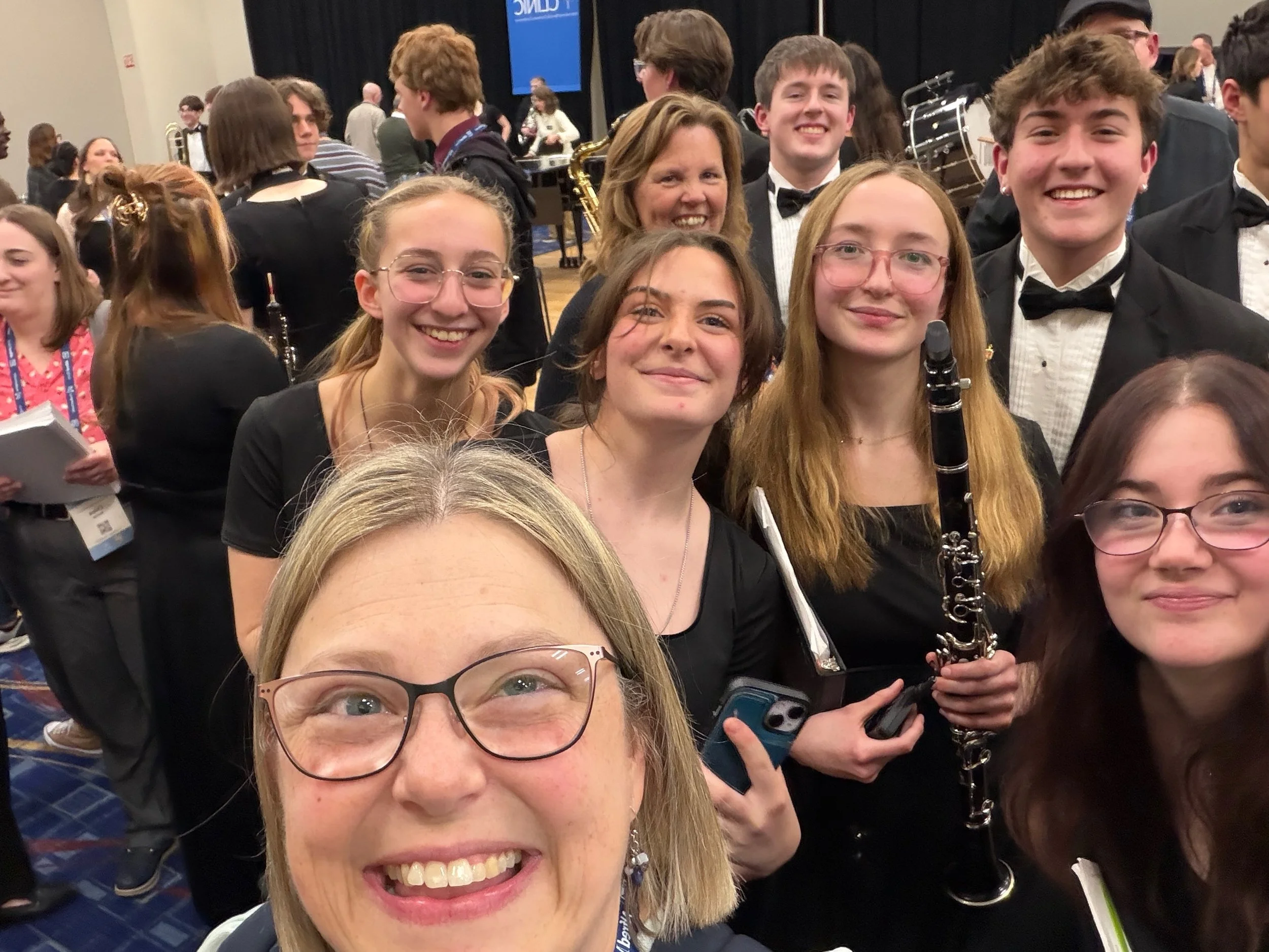 Group of young people, mostly women, dressed in formal attire, gathered at a music event with a woman in the foreground smiling and taking a selfie. A musician with a clarinet is visible in the background.