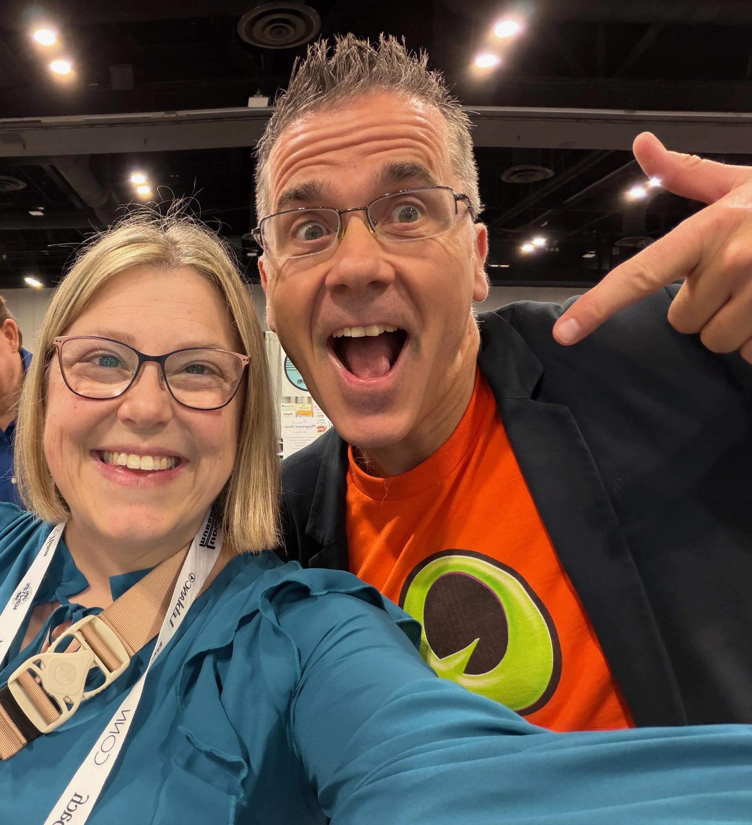 Close-up of two smiling individuals at an indoor event, with the man on the right making a peace sign. The woman on the left has blonde hair and wears glasses, while the man has spiked hair, glasses, and a colorful shirt with a large design. The back