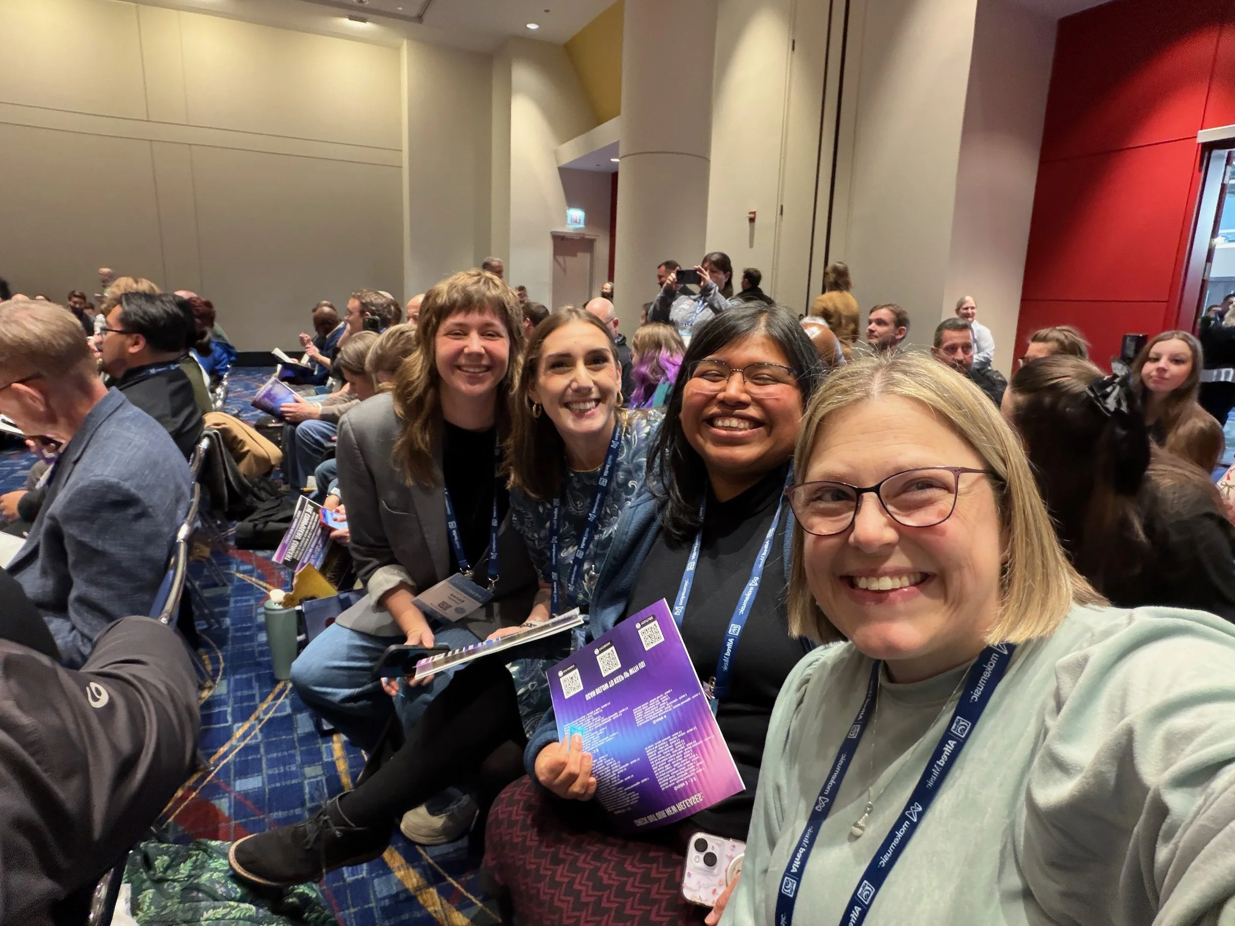 A group of smiling people at a conference or event, some holding pamphlets, in a room with beige walls and a colorful carpet.