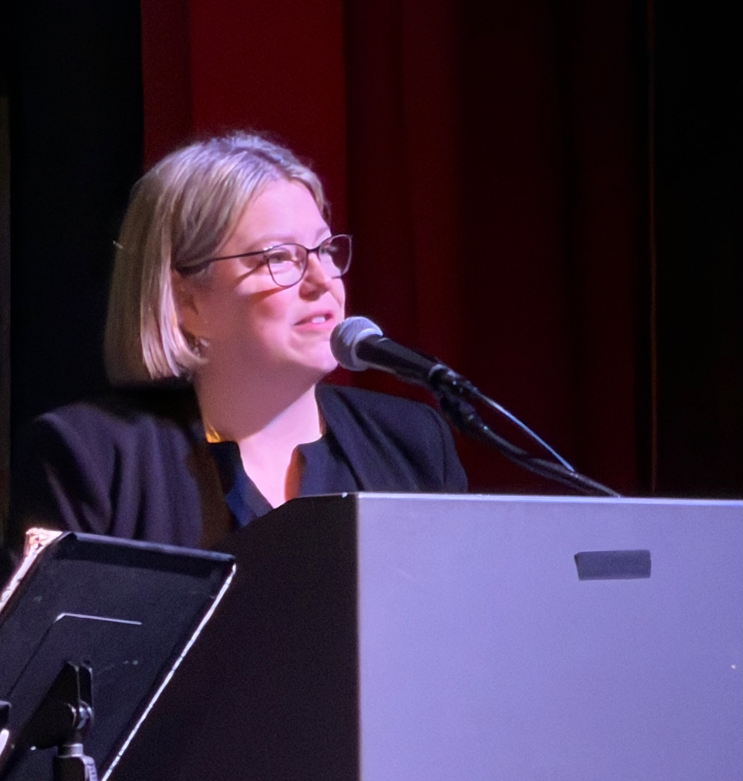 A woman with blonde hair and glasses speaking into a microphone on a stage with a dark background.