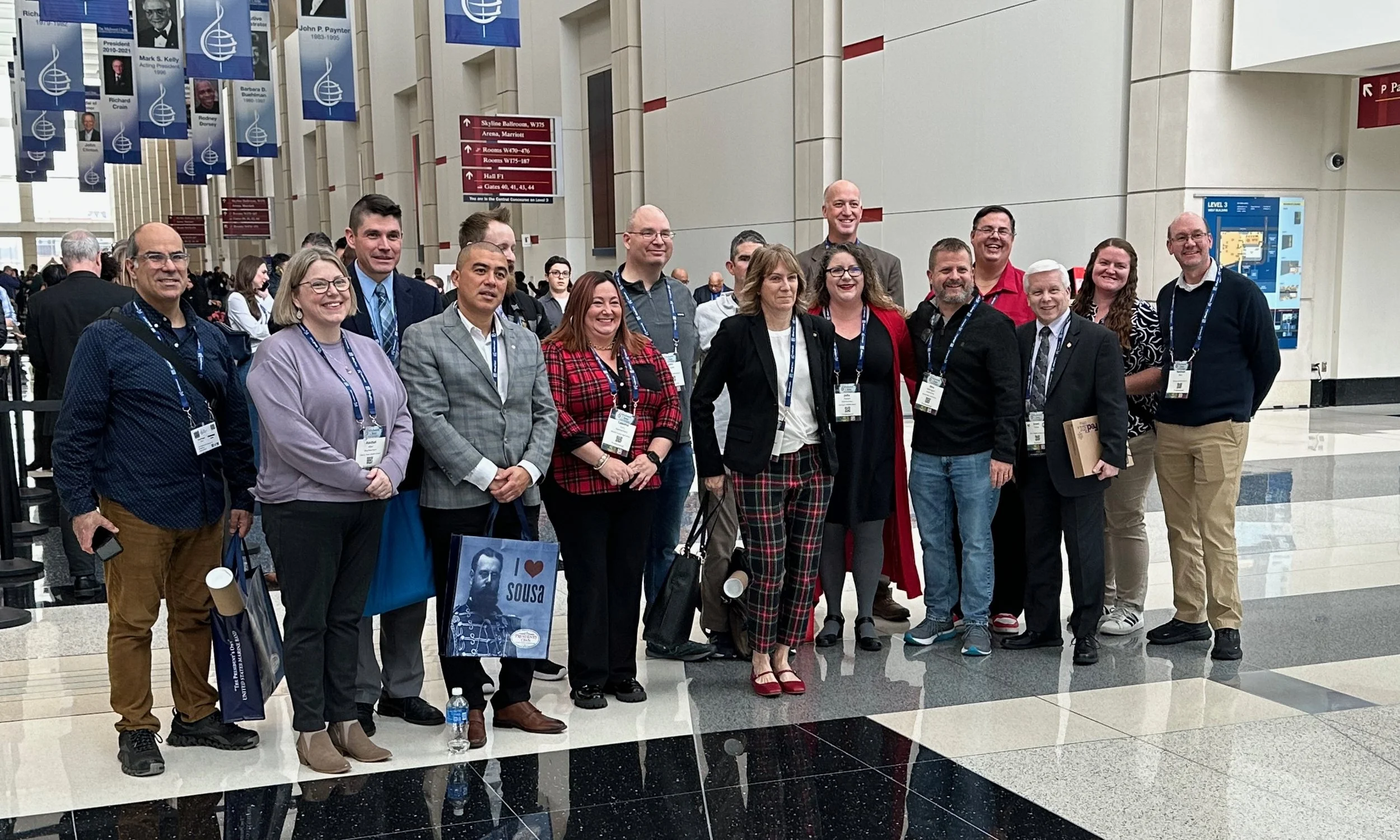 Group of diverse people standing indoors at a conference or event, smiling and posing for a photo. They are wearing name tags and some are holding bags and documents. In the background, banners and signs with directional information are visible.