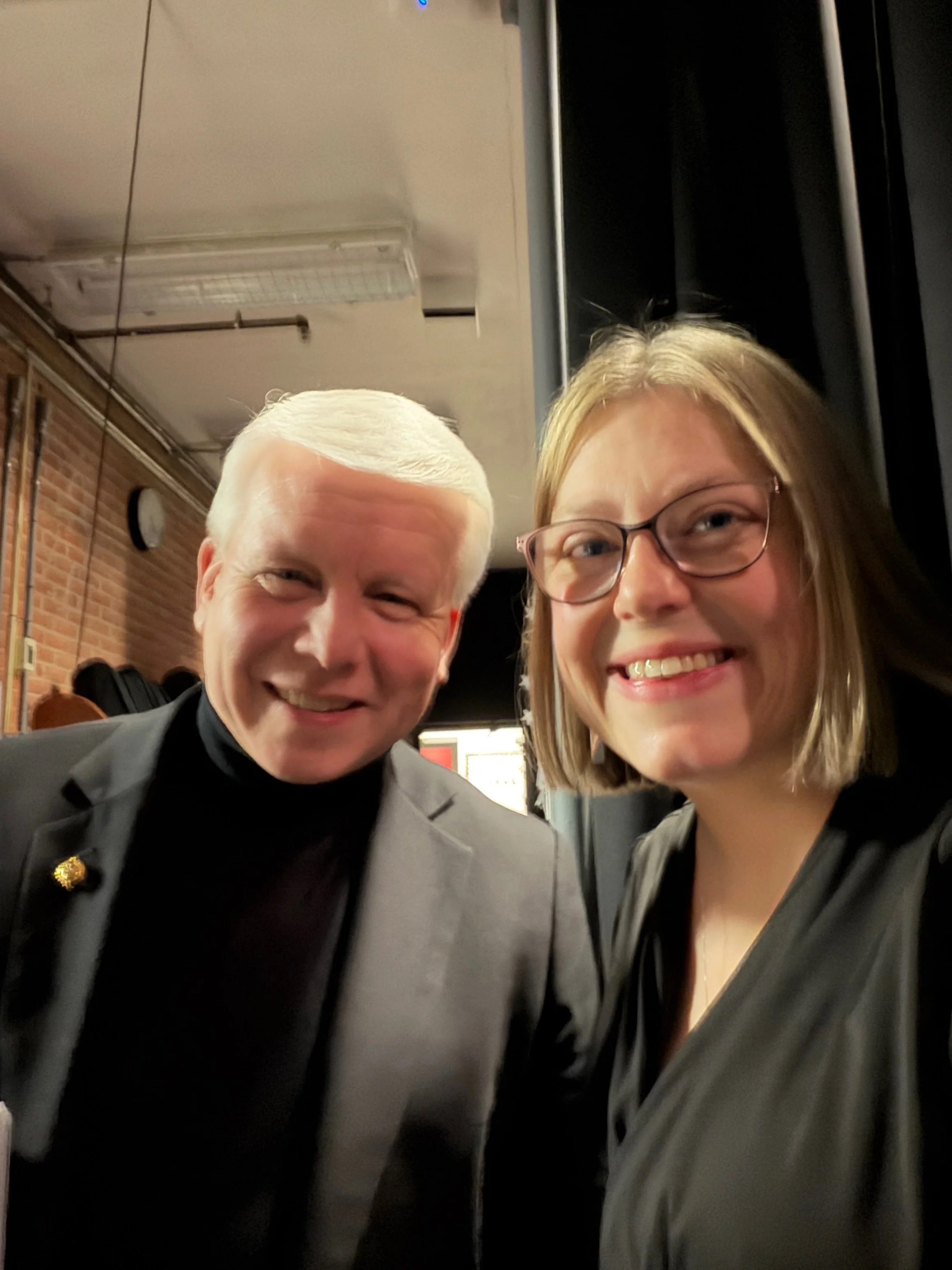 Two smiling people posing for a selfie in an indoor location with brick walls visible in the background.