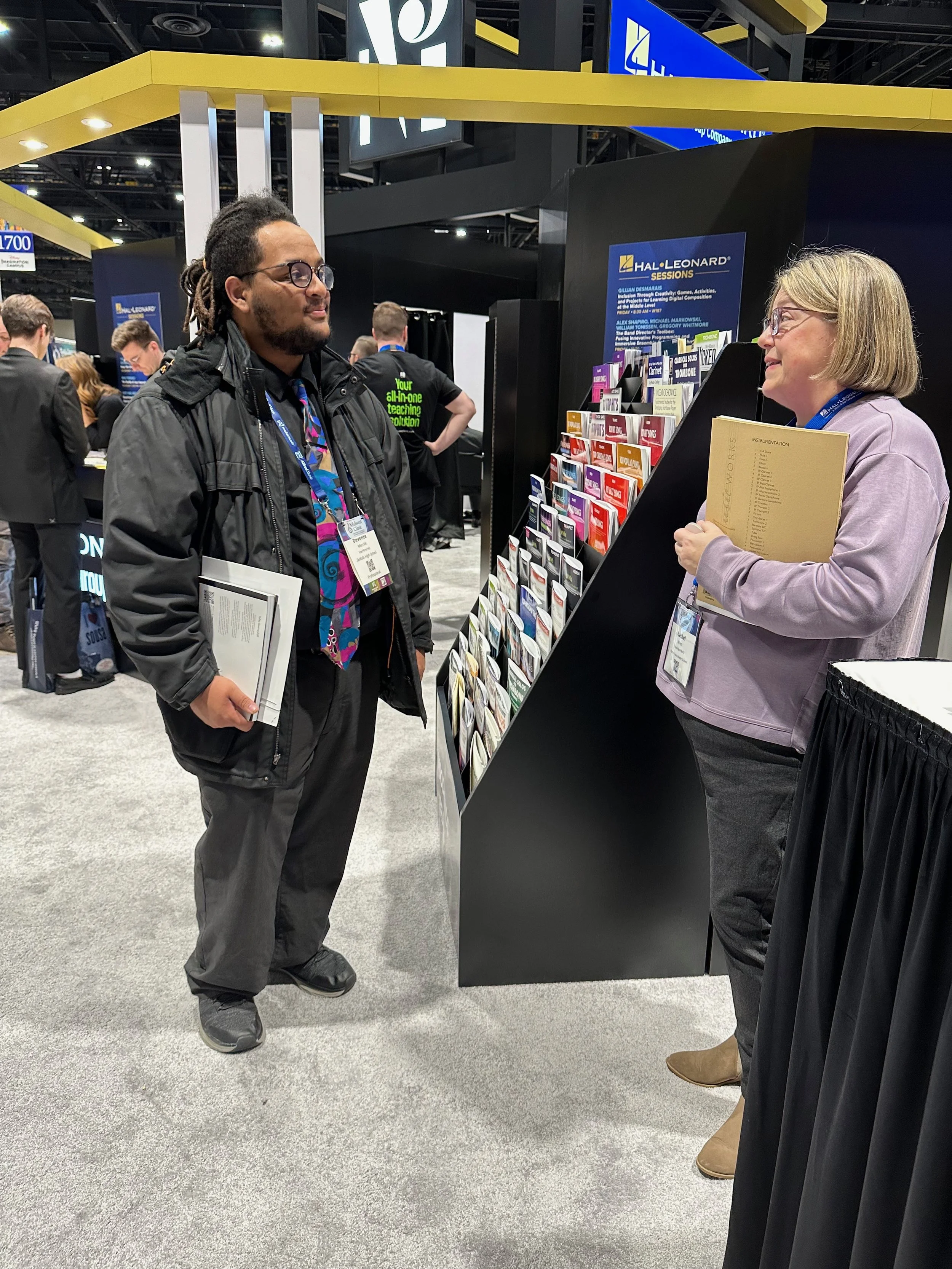 Two people, a man and a woman, are having a conversation at a trade show or conference booth, with the man holding a notebook. They are standing in front of a display stand with various pamphlets or brochures. The background shows other attendees and