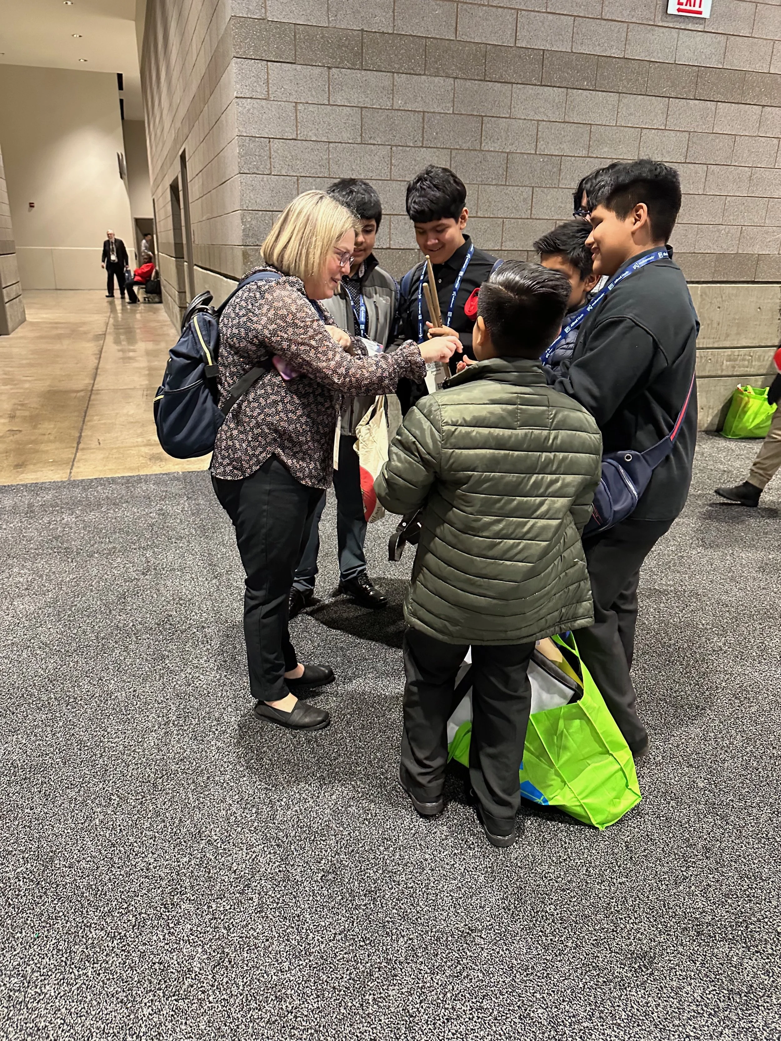 A group of six people, four boys and two adults, gathered together in a semi-circle in a hallway, looking at something held by the woman with blonde hair and a patterned blouse, while the boys wear dark clothing and lanyards. One boy has a green and 