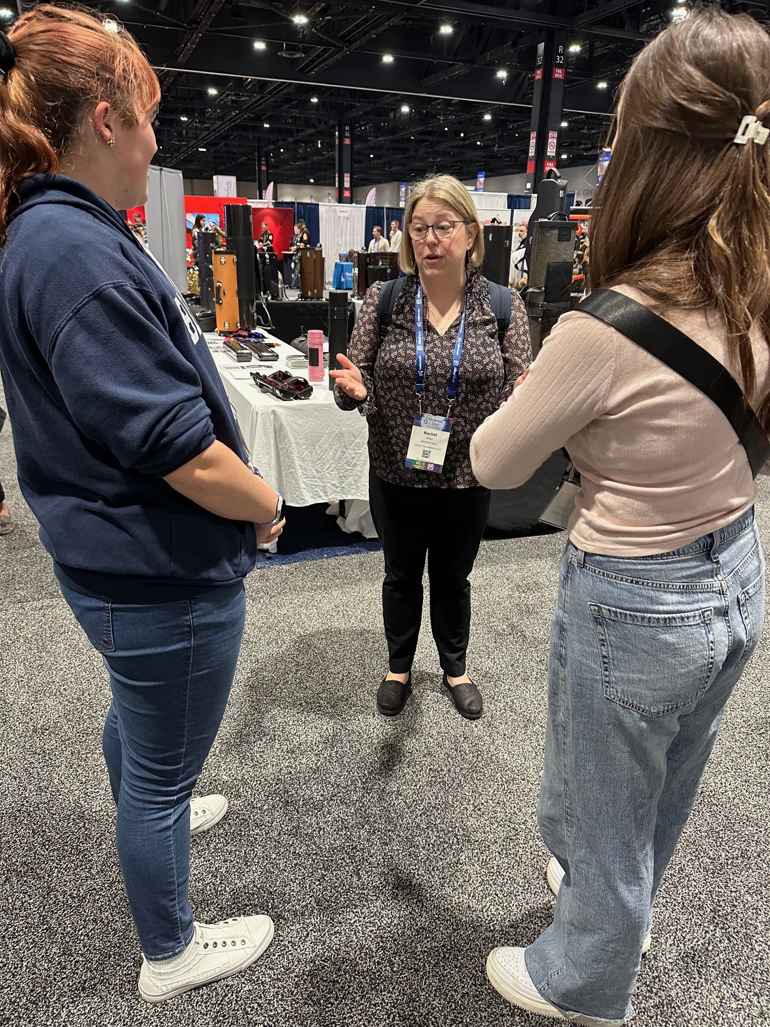 Three women are standing and talking at an indoor event or trade show. The middle woman is speaking and gesturing with her hands, while the other two women listen. There are booths and displays in the background.