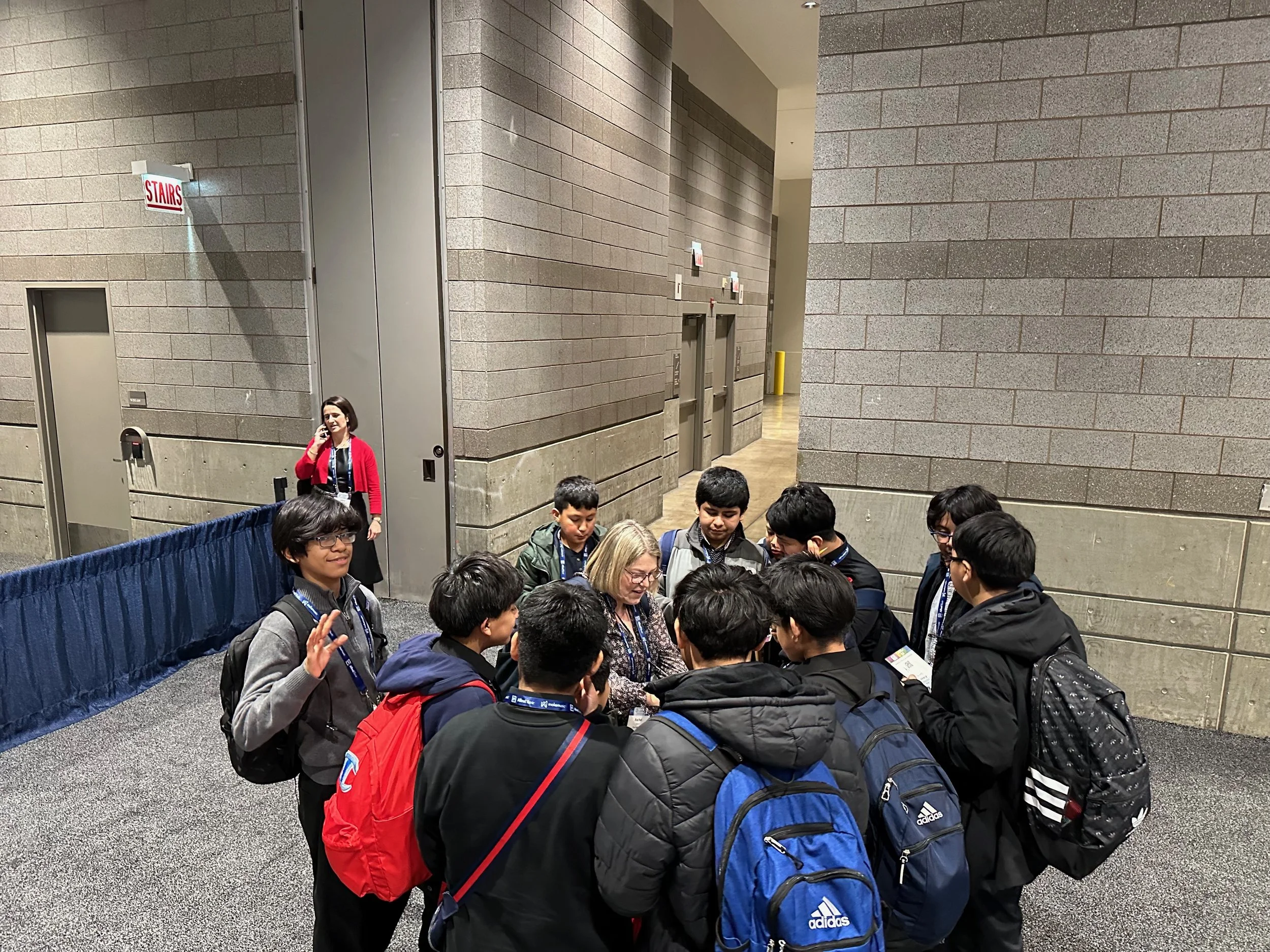 A group of young people, possibly students, standing in line and engaged in conversation at an indoor location with gray brick walls. One woman dressed in red is talking on a cellphone near a wall with a sign that reads 'Stairs'. Some people are carr