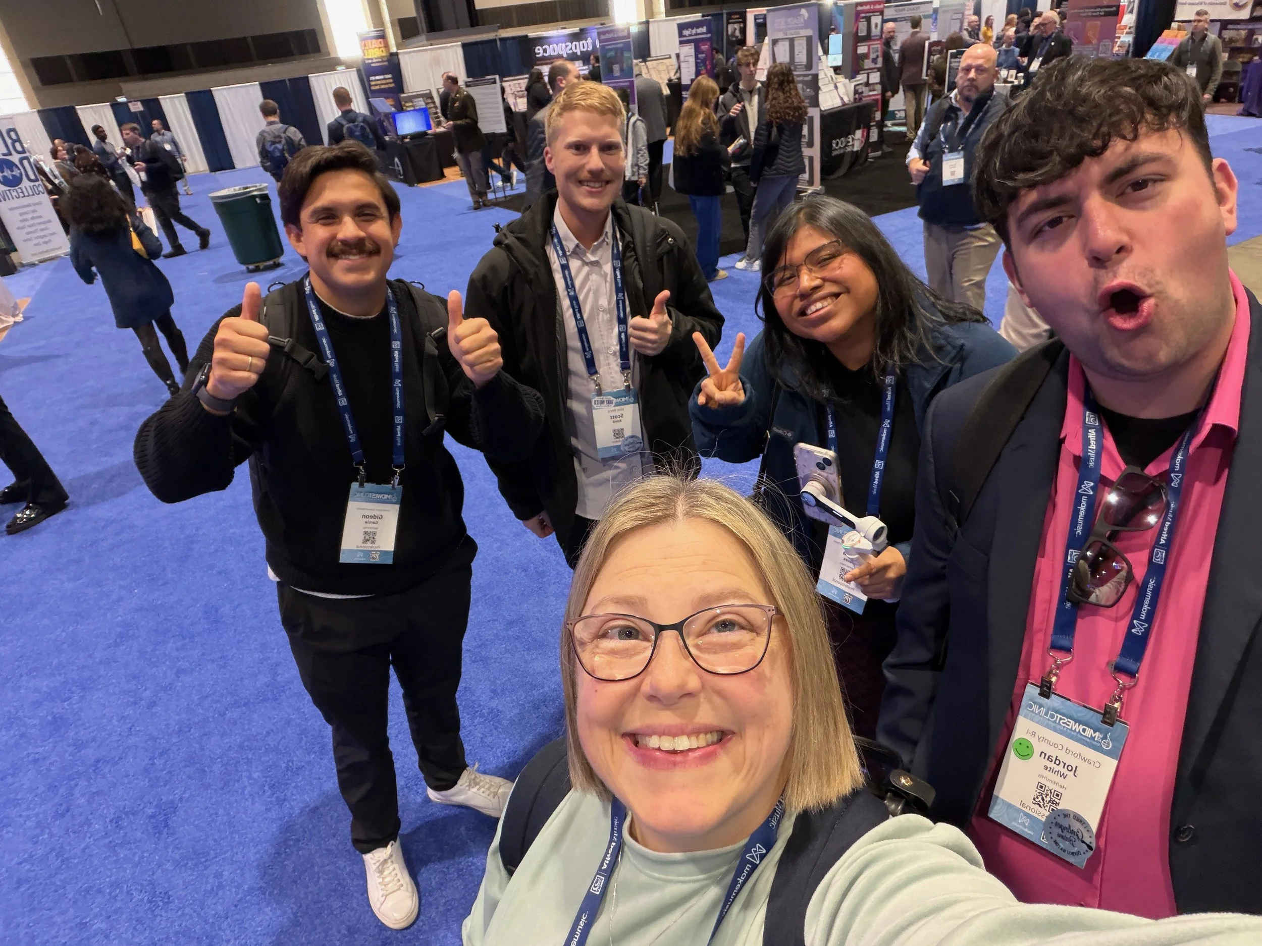 Four people taking a selfie at a conference or event, with a blue carpeted floor and exhibition booths in the background. They are smiling and making gestures such as thumbs-up and peace sign.