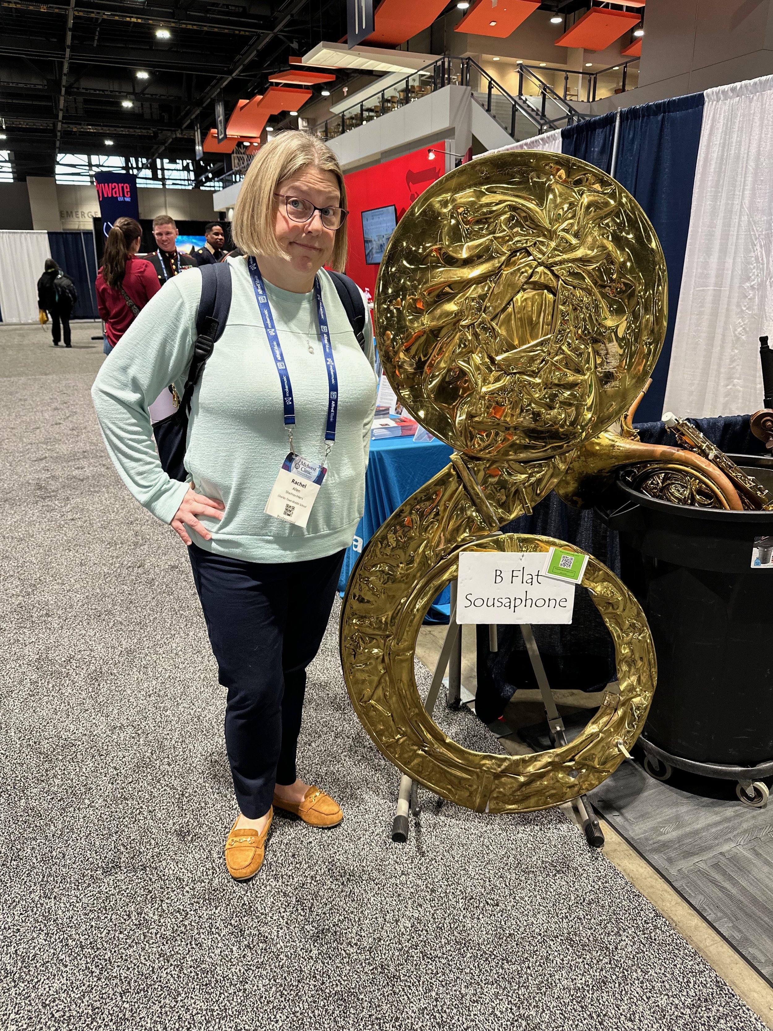 A woman with shoulder-length blonde hair, glasses, and a light green sweater stands beside a large, shiny, gold-colored sousaphone at an indoor event. She has a name badge around her neck that reads 'Rachel.' There are people and booths in the backgr