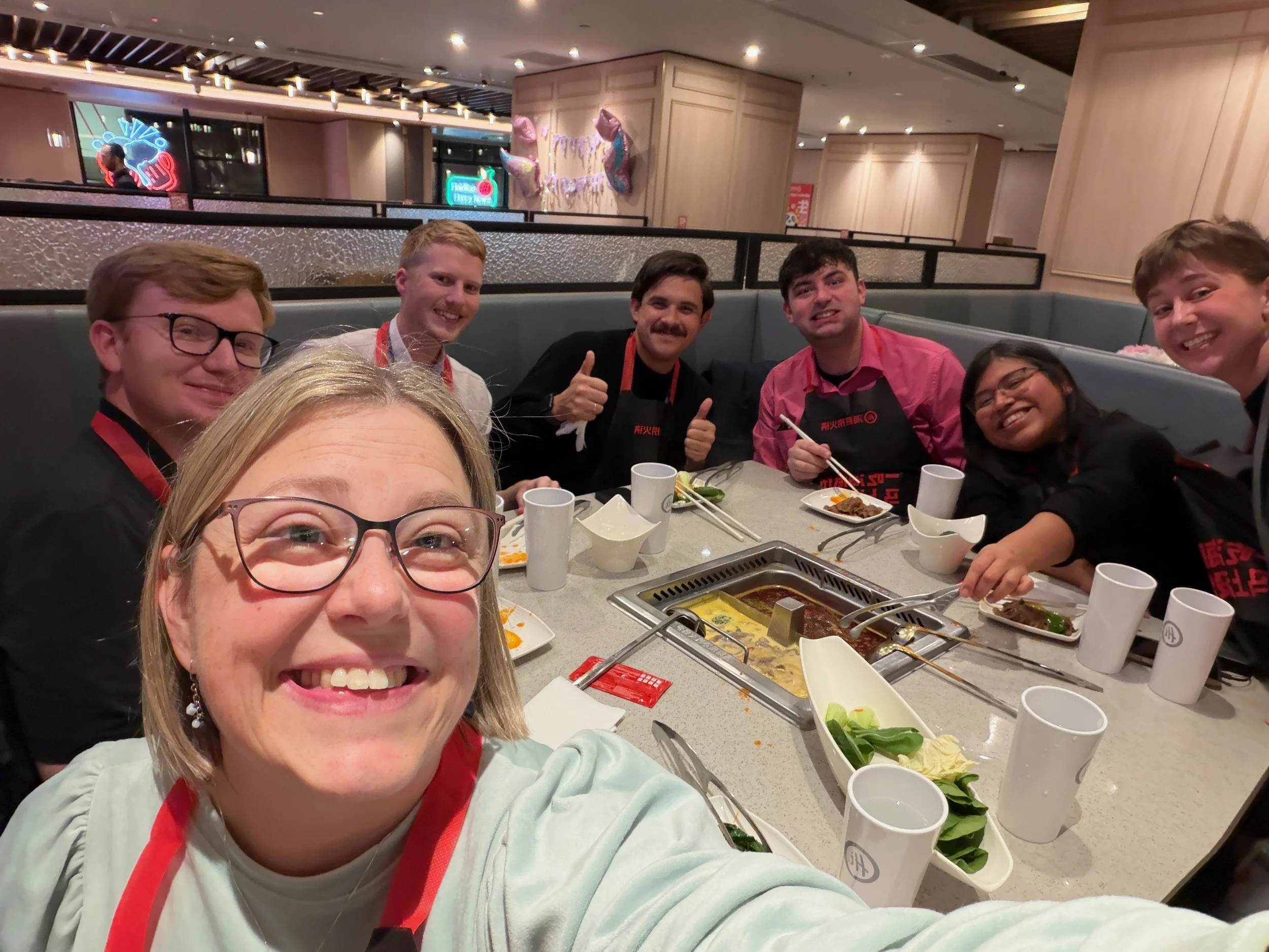A group of seven people sitting around a table at a restaurant, smiling and taking a selfie. The table has plates with food, cups, and chopsticks, indicating they are enjoying a meal together.