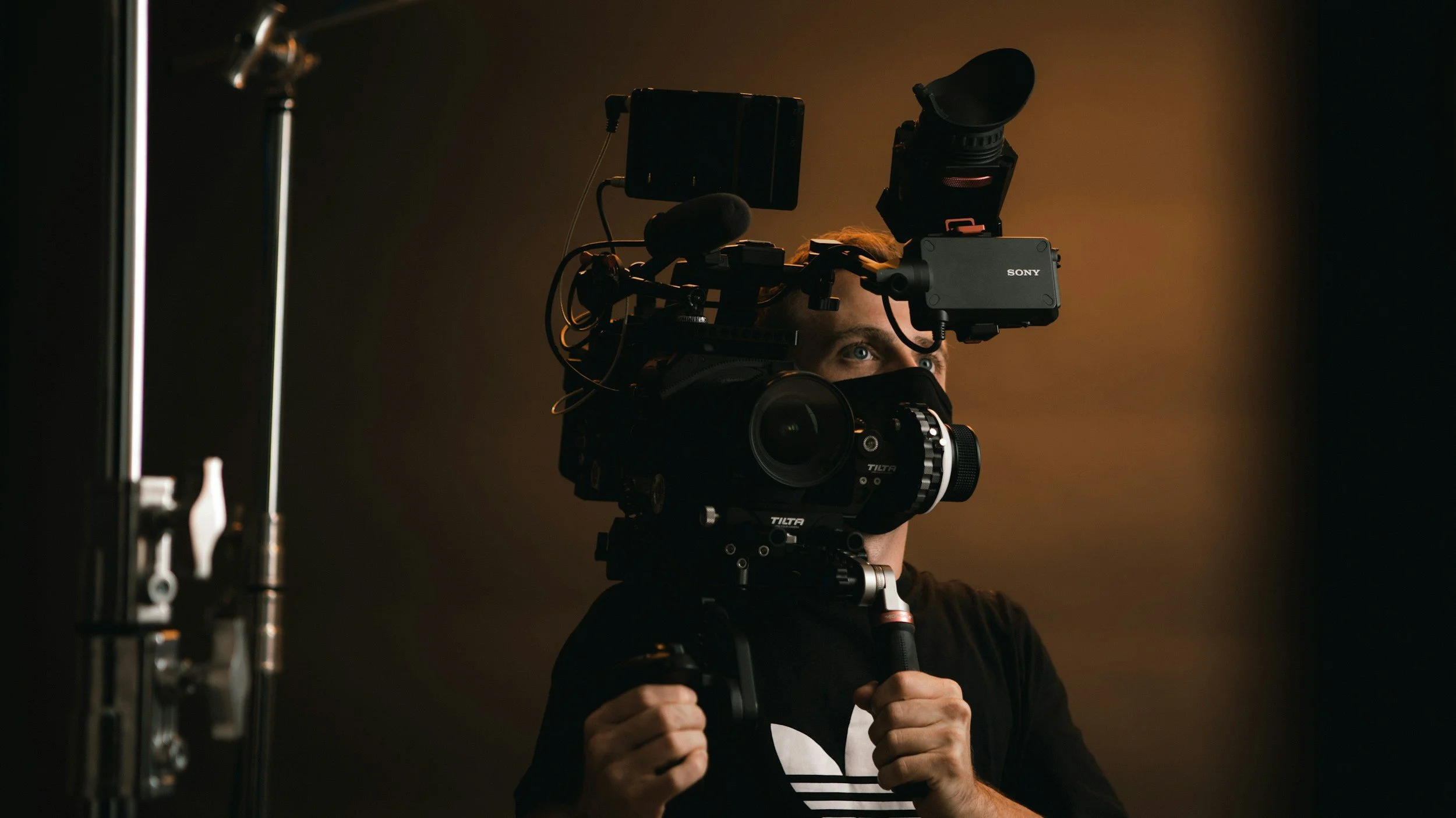 A person holding a camera rig on their shoulder, with various mounted equipment including a monitor and microphone, in a dark studio setting.