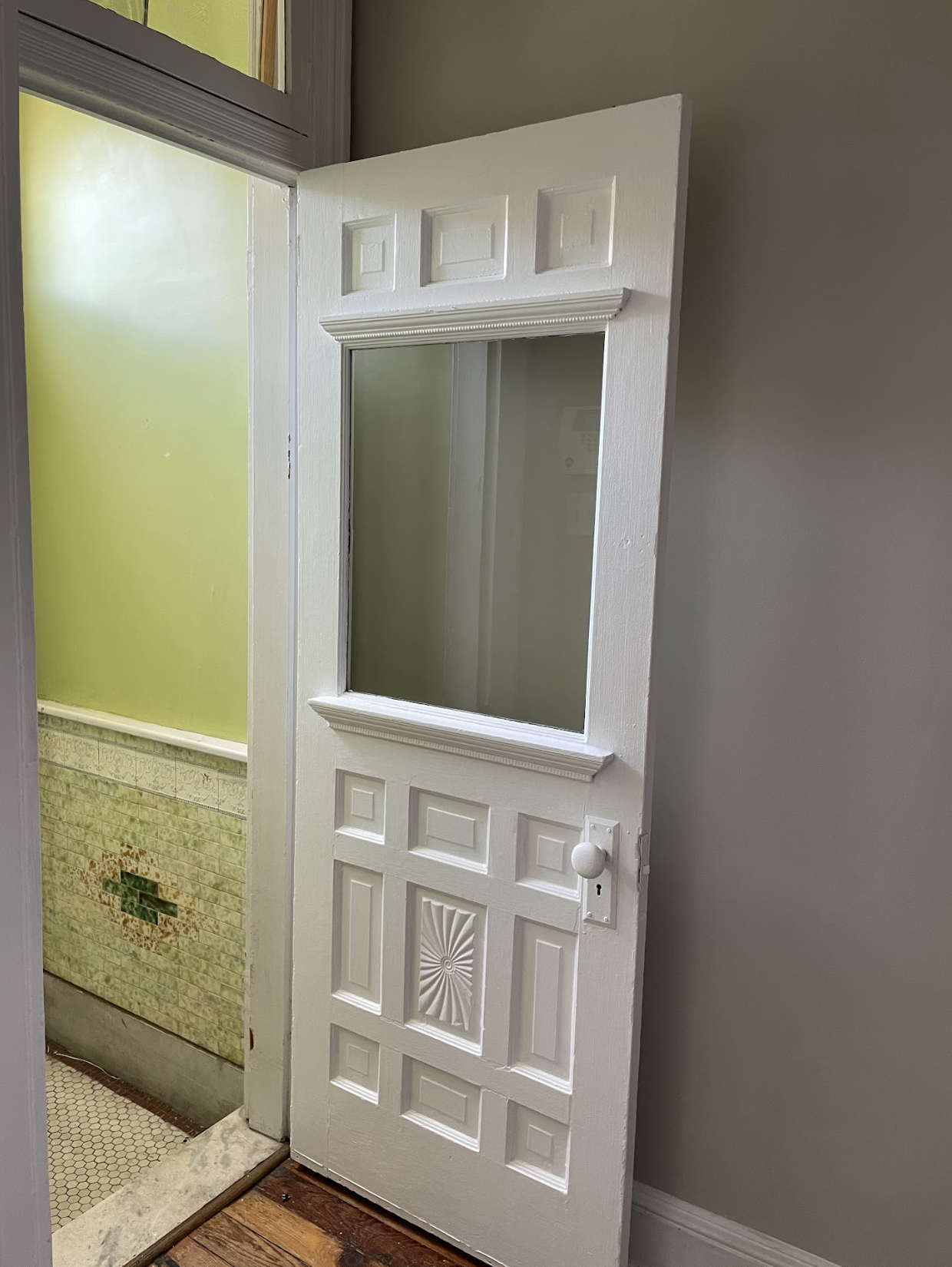Open white interior door with decorative panels, glass window, and round doorknob in a room with green tiled walls and tile floor.