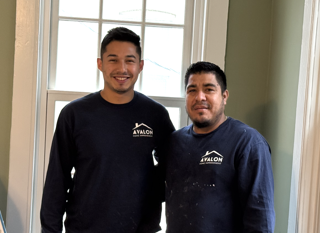 Two men wearing Avalon Home Improvement shirts standing indoors in front of a window.