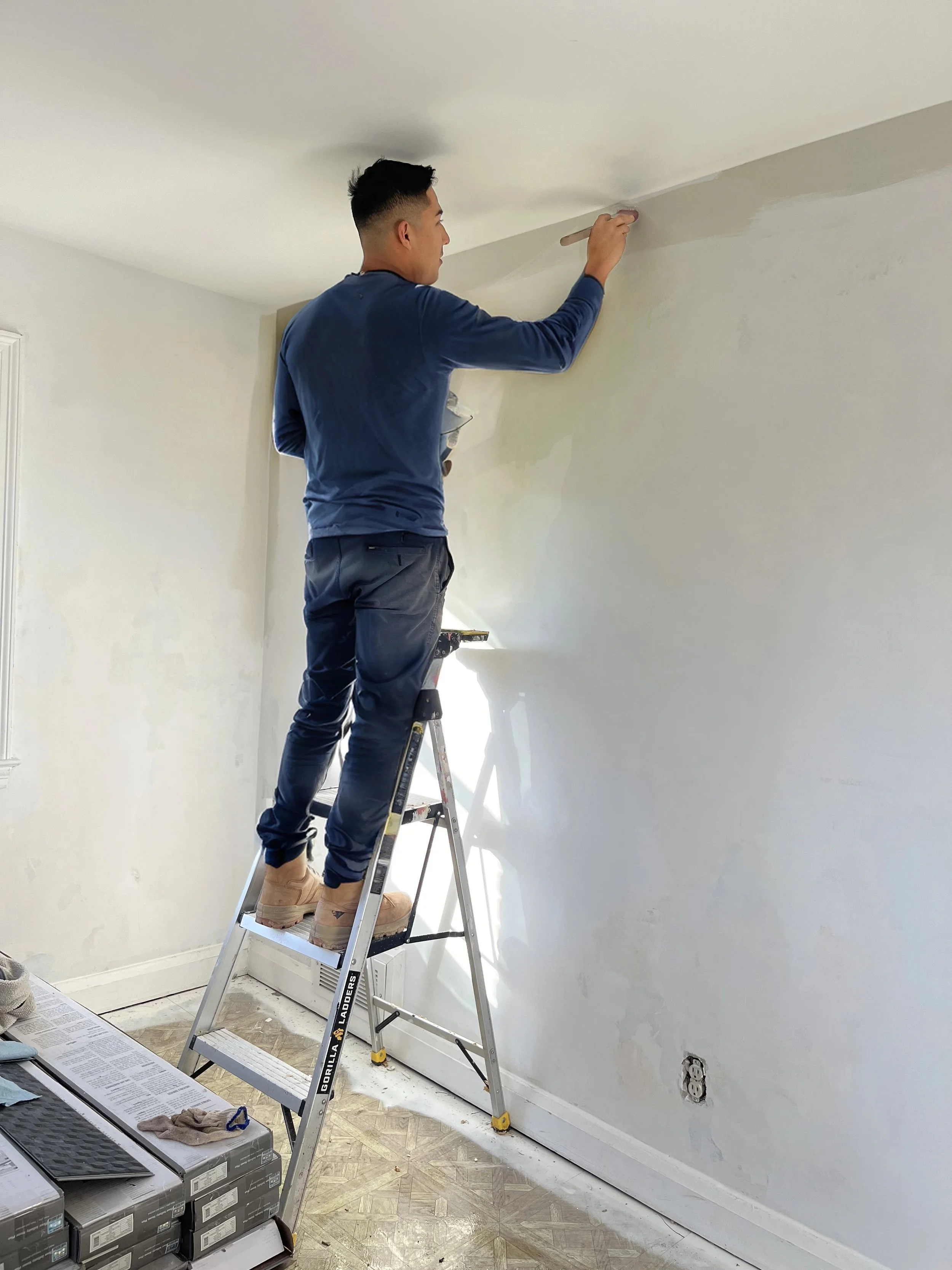 Man in blue shirt painting tan wall while standing on ladder