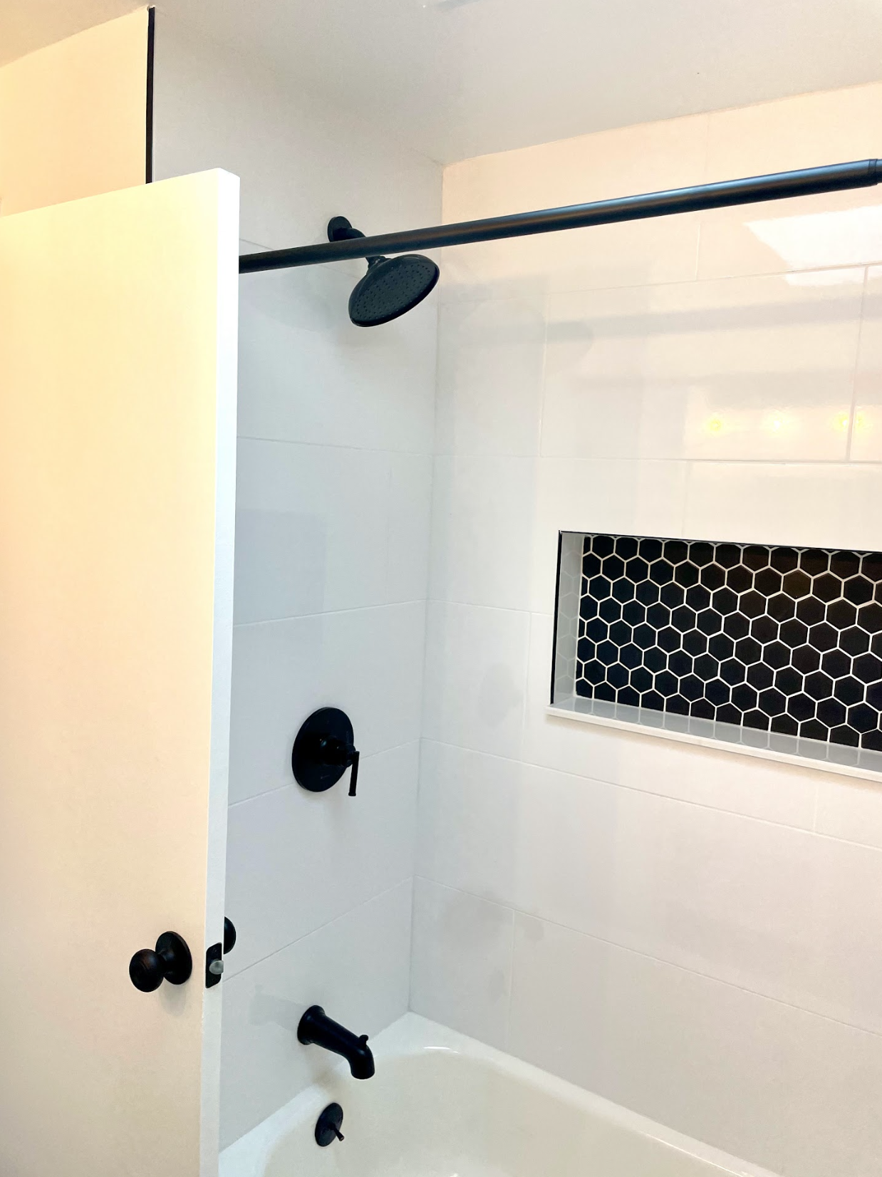 Modern bathroom with a white tiled tub and shower area, featuring black fixtures including a showerhead, faucet, and handles. The shower has a honeycomb-patterned black tile niche.