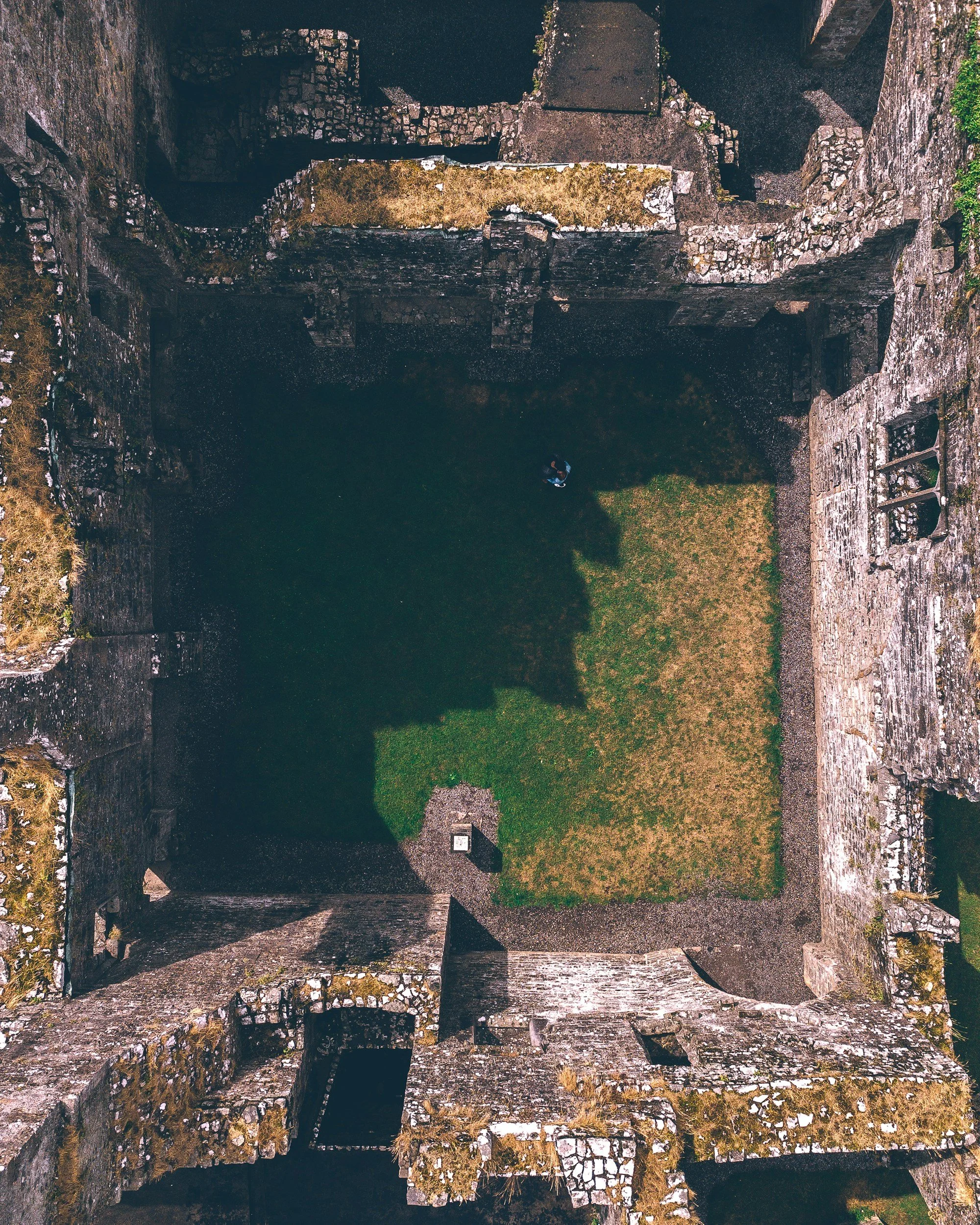 Aerial view of an ancient stone structure with moss-covered walls surrounding a grassy courtyard, with one person walking inside.
