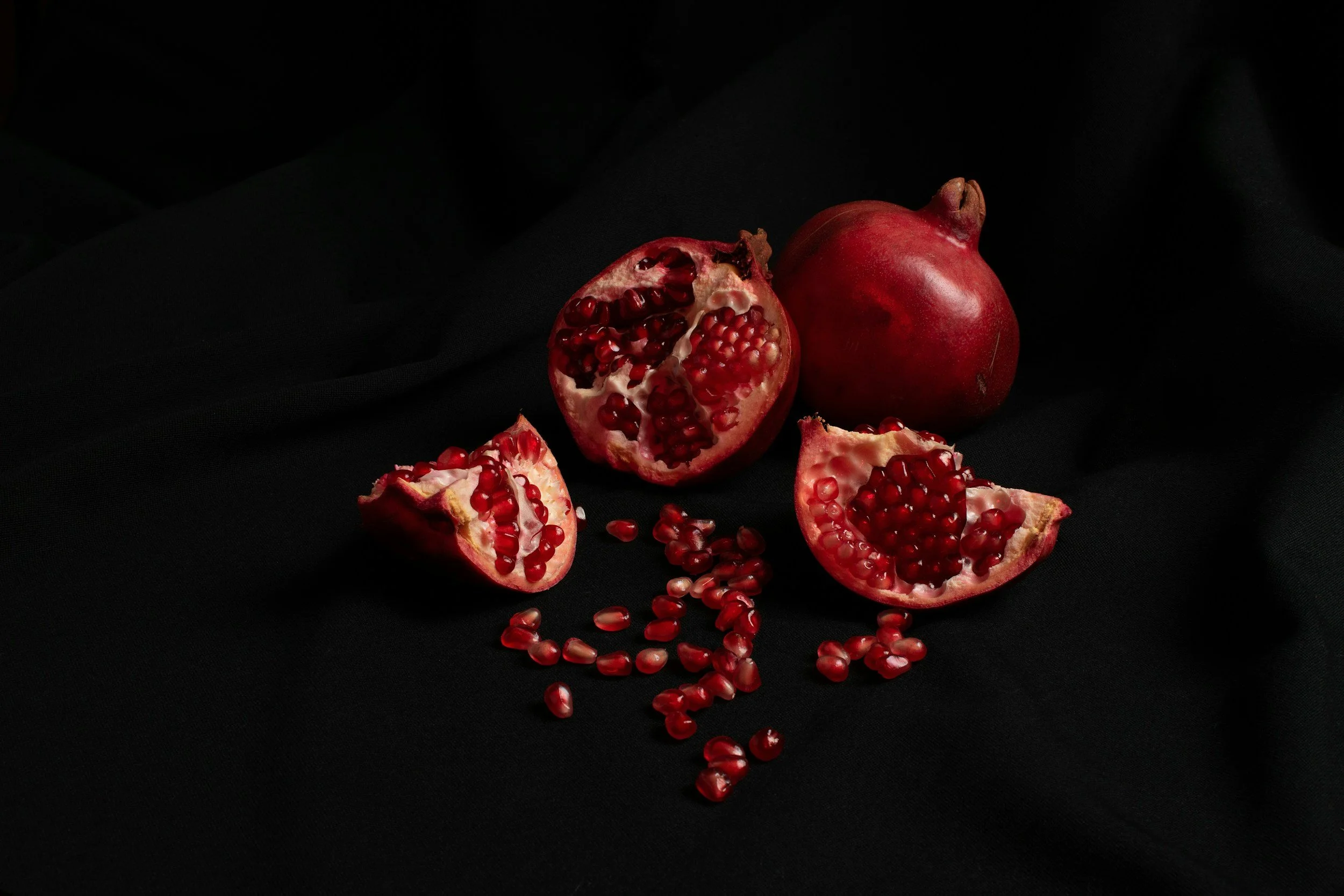 A whole pomegranate fruit, a halved pomegranate, and two pomegranate wedges with seeds scattered on a black fabric background.