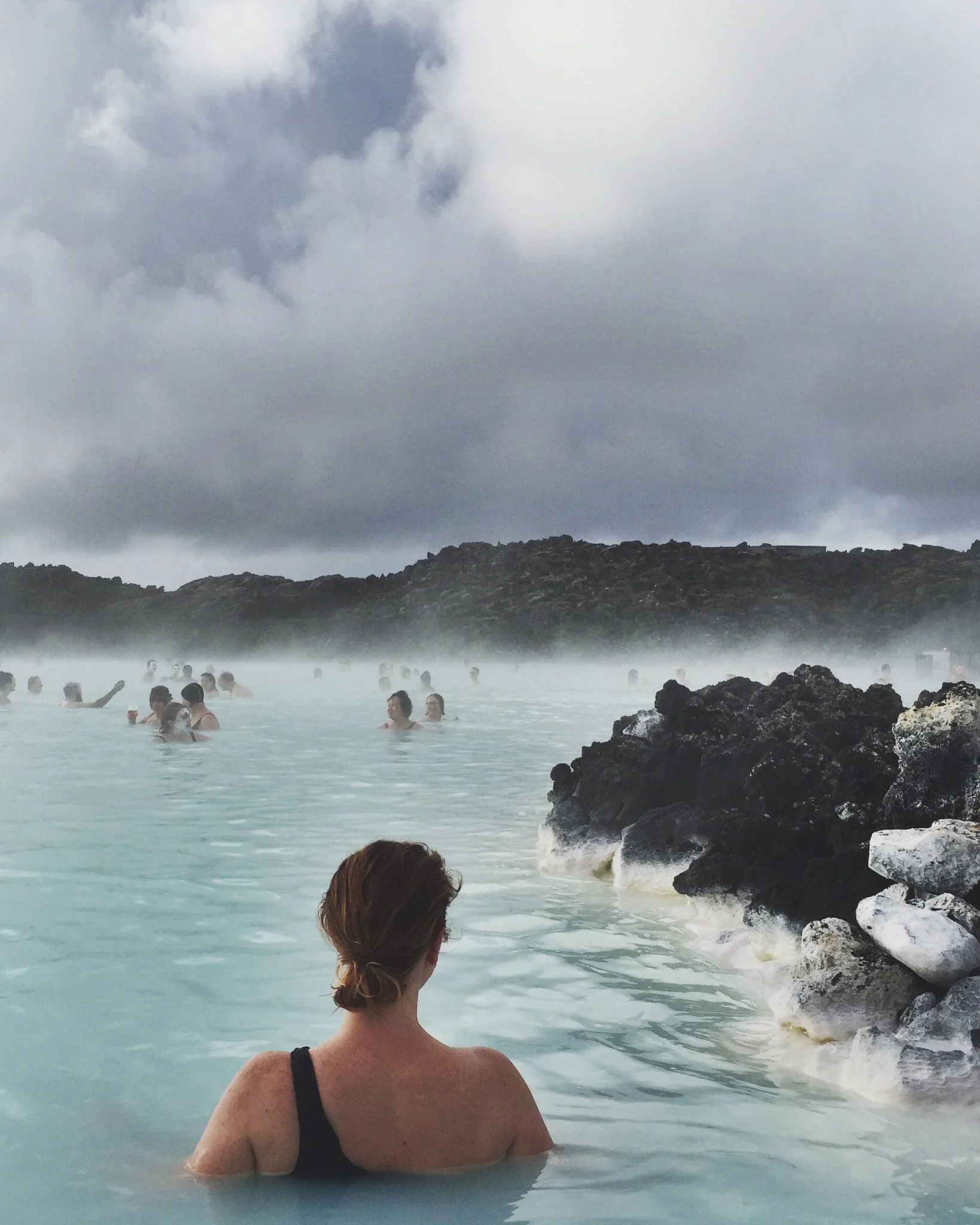 People relaxing in a geothermal hot spring pool with dark volcanic rocks, cloudy sky, and steam rising from the water.