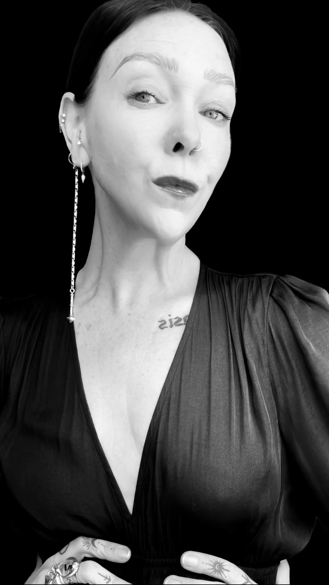 A woman with short dark hair and light-colored eyes wearing a black dress with pleats, multiple earrings, a septum piercing, and tattoos on her fingers and chest. She is posing against a black background.
