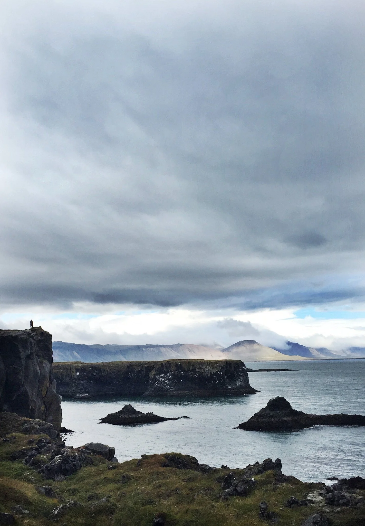 A rugged coastline with a person standing on the edge of a cliff, surrounded by dark clouds, distant mountains, and a body of water.